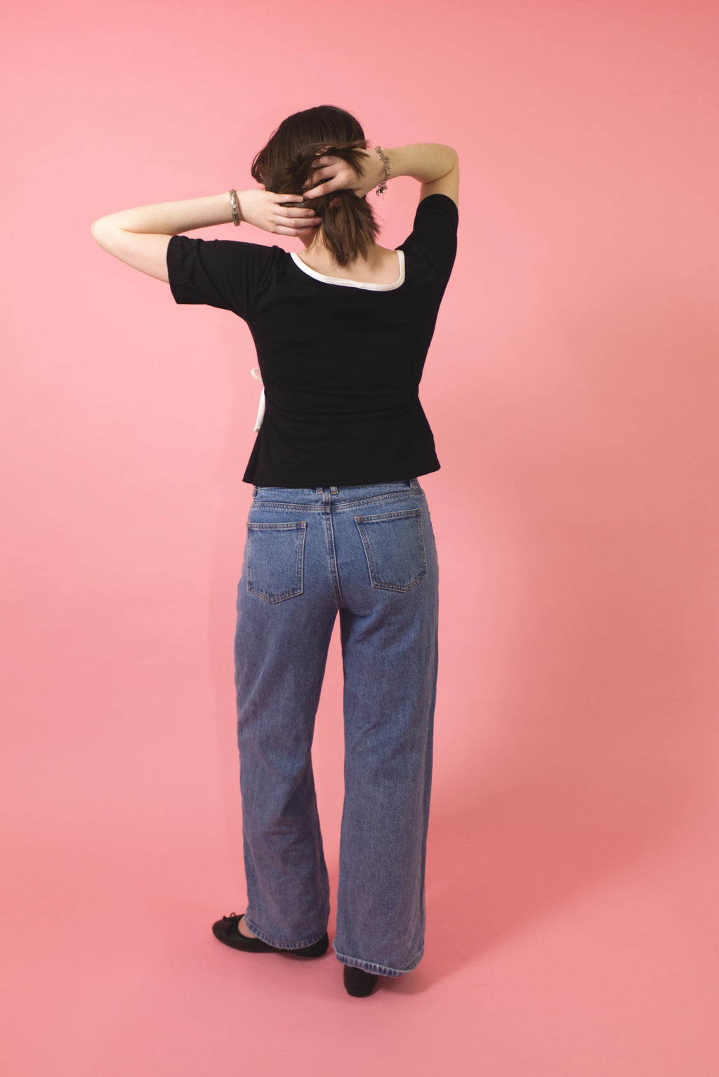 A woman viewed from behind wearing a black short-sleeve top with a white scoop neckline and wide-leg blue jeans, standing against a solid pink background.
