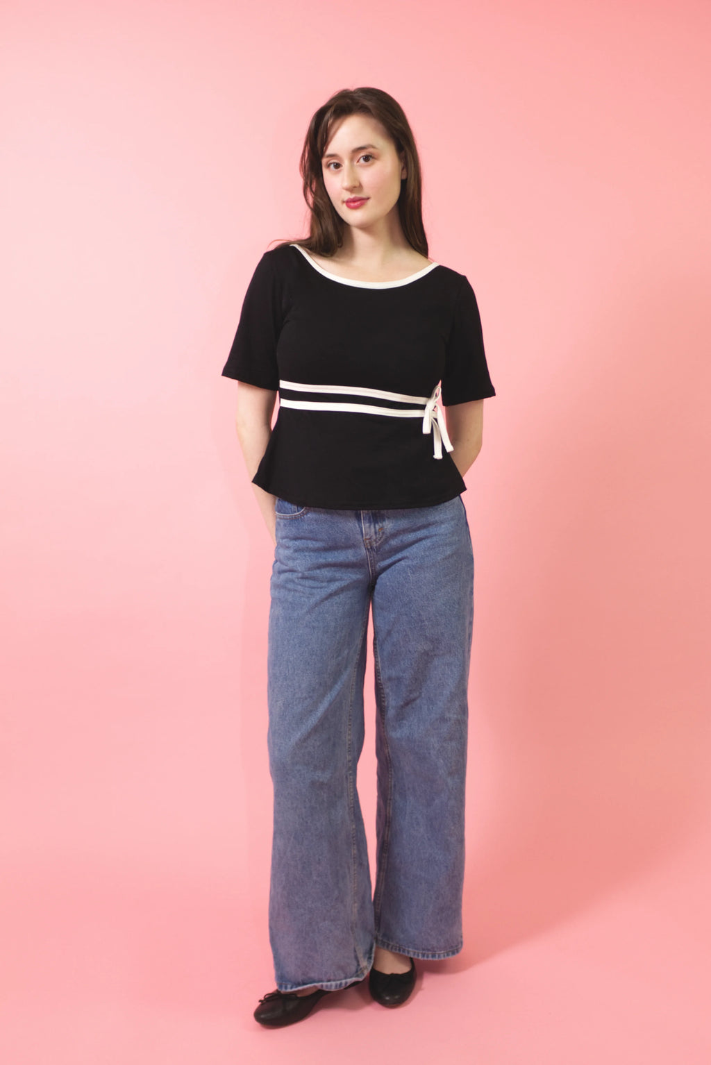 A woman modeling a black short-sleeve top with white trim and a white horizontal stripe detail, paired with wide-leg blue jeans, standing against a solid pink background.