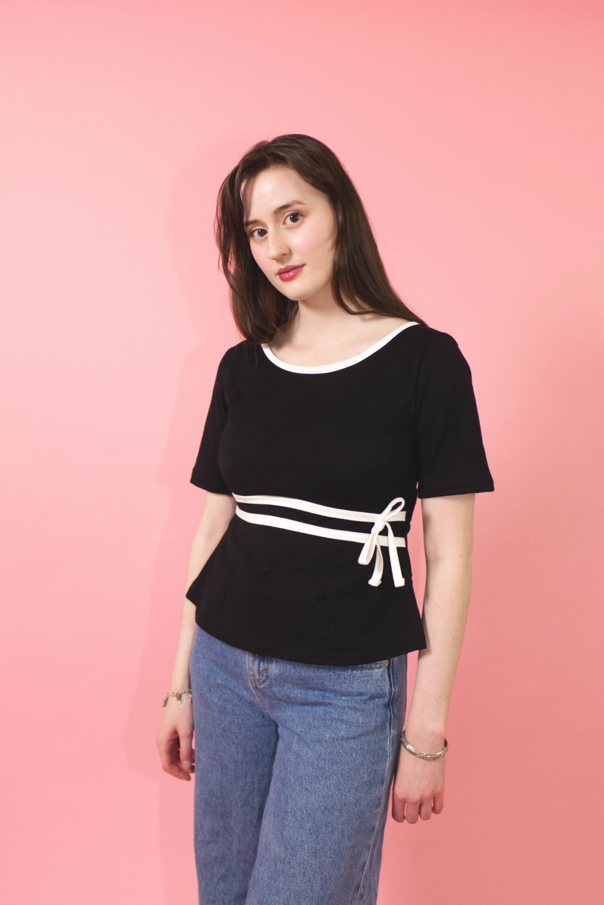 A woman modeling a black short-sleeve top with a wide white neckline, a horizontal white stripe across the waist, and a white bow detail on the right side, paired with blue jeans against a solid pink background.