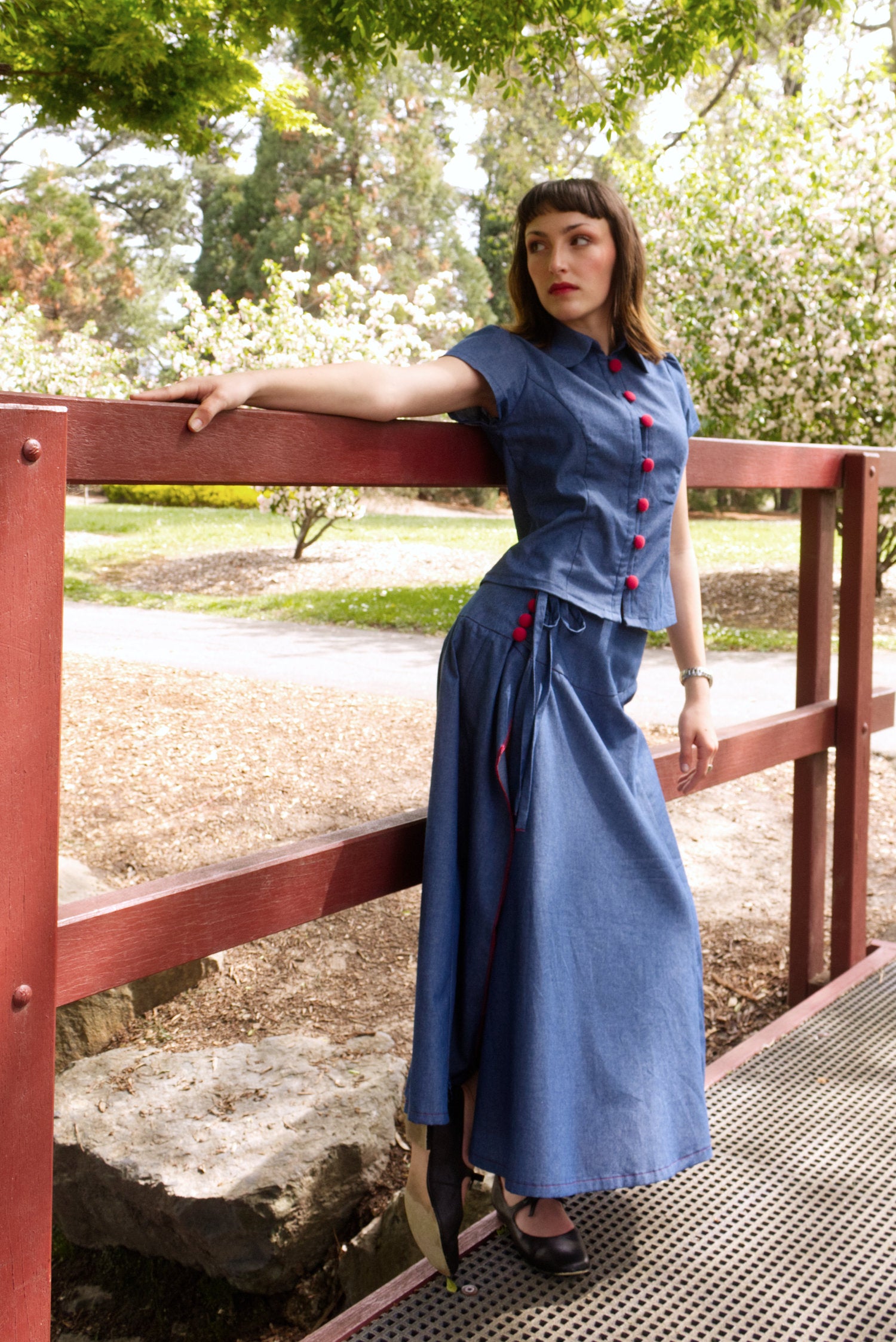 Young woman in a matching blue chambray cap sleeve princess line blouse and asymmetrical maxi warp skirt with a split, both with red buttons, on a bridge in a park