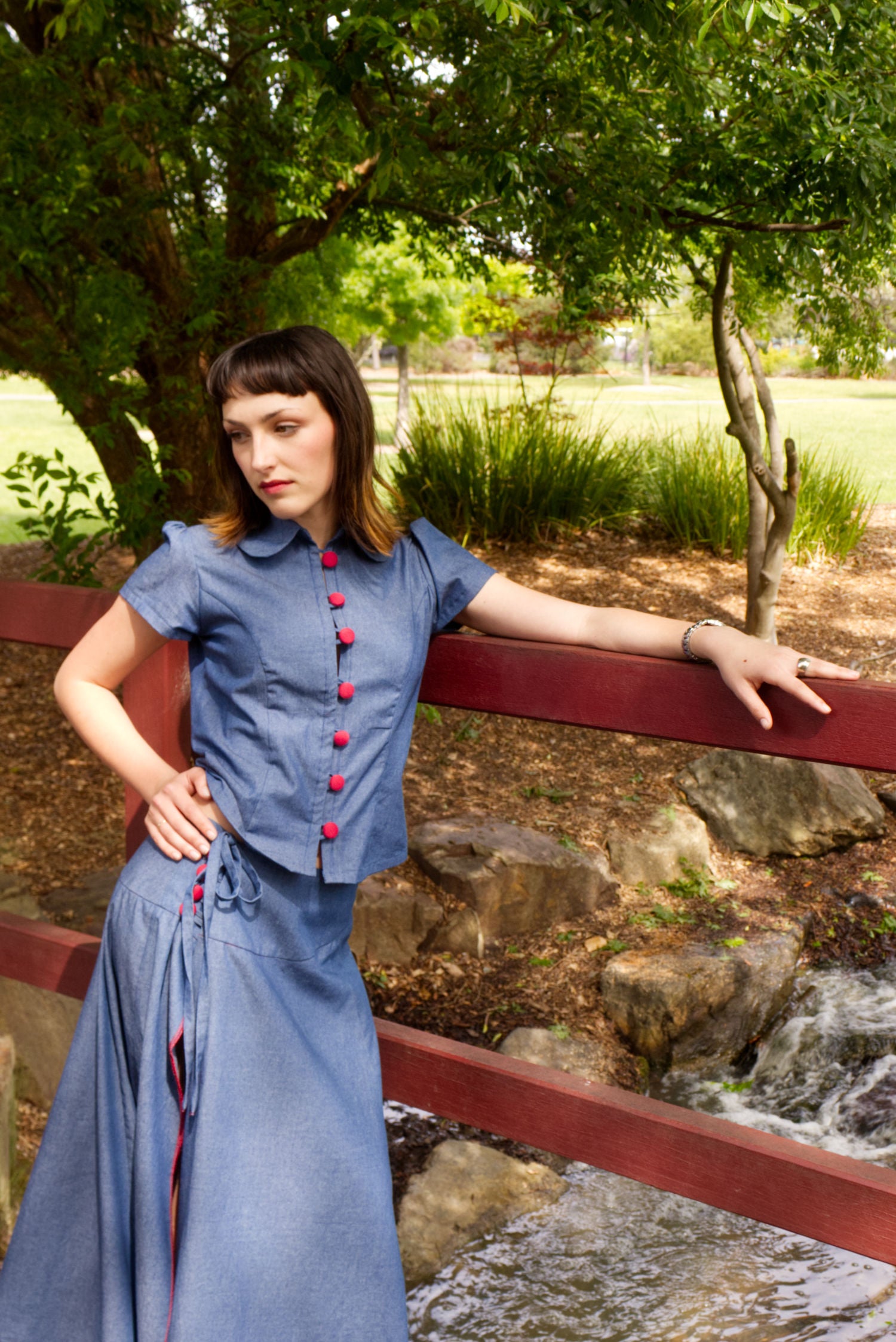 Young woman in a matching blue chambray cap sleeve princess line blouse and asymmetrical maxi warp skirt with a split, both with red buttons, on a bridge in a park