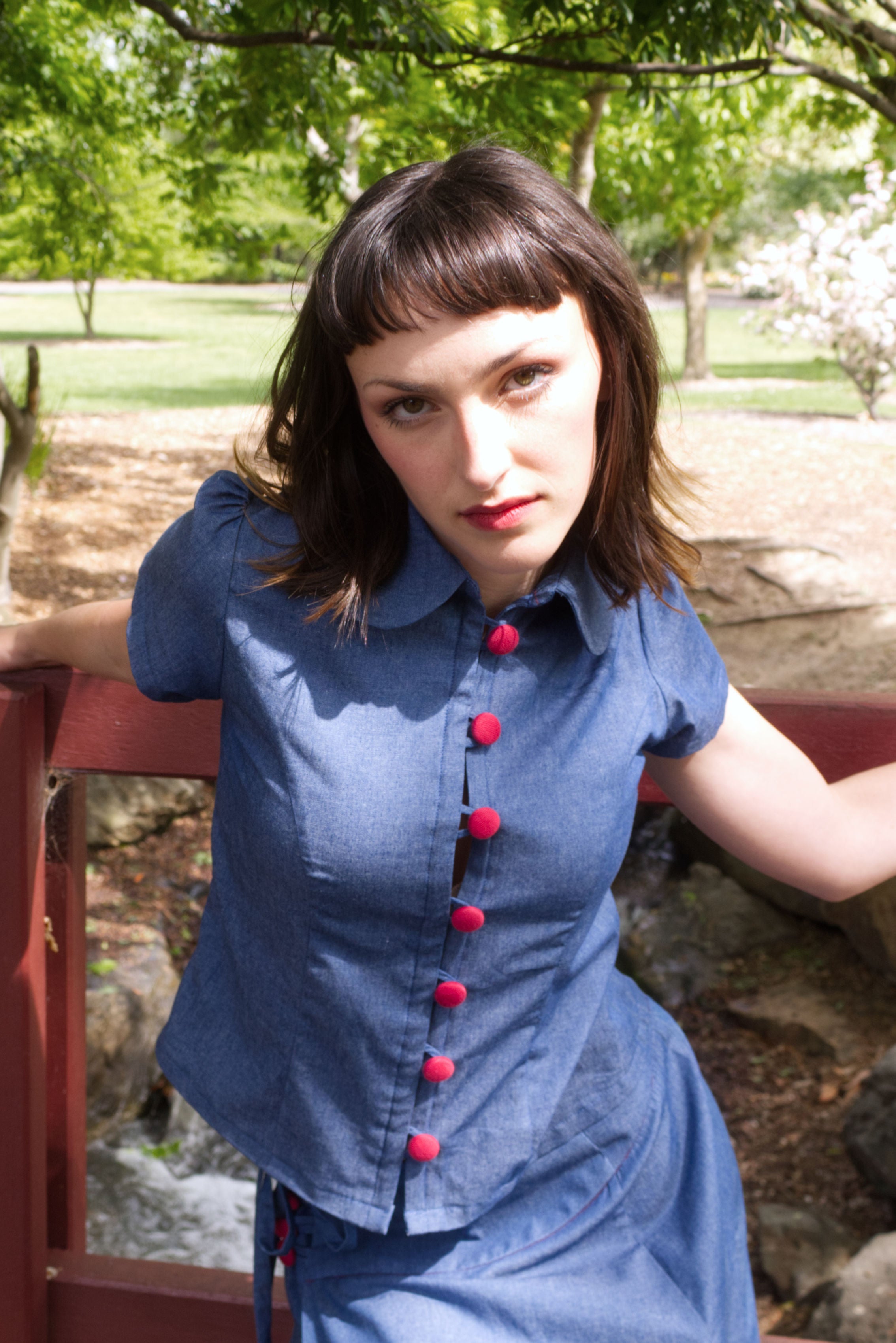 Young woman in a matching blue chambray cap sleeve princess line blouse and asymmetrical maxi warp skirt with a split, both with red buttons, on a bridge in a park