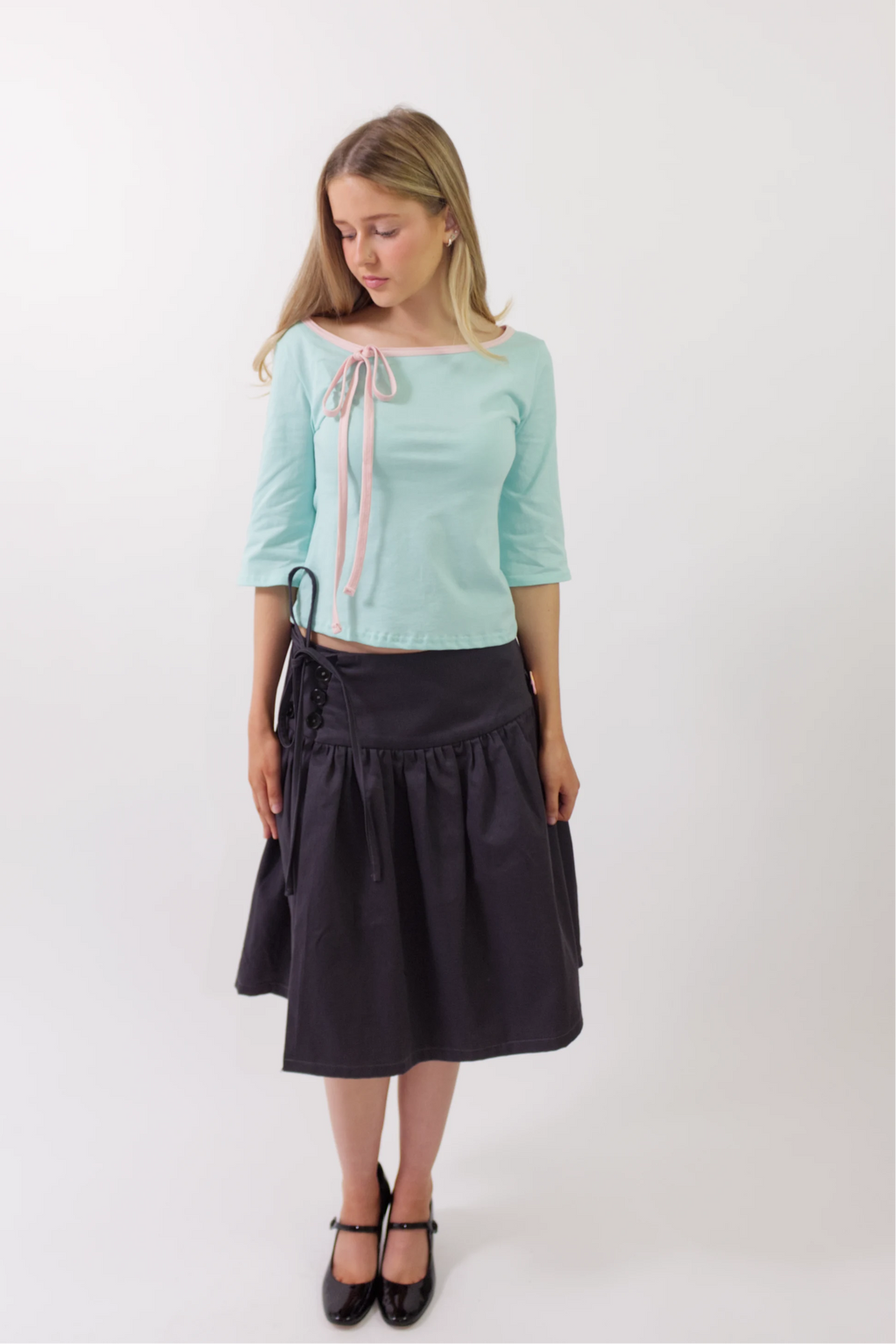 A woman modeling a light mint green three-quarter sleeve top with a pink and white striped tie detail at the neckline, paired with a dark navy midi skirt and black Mary Jane shoes, against a plain white studio background.
