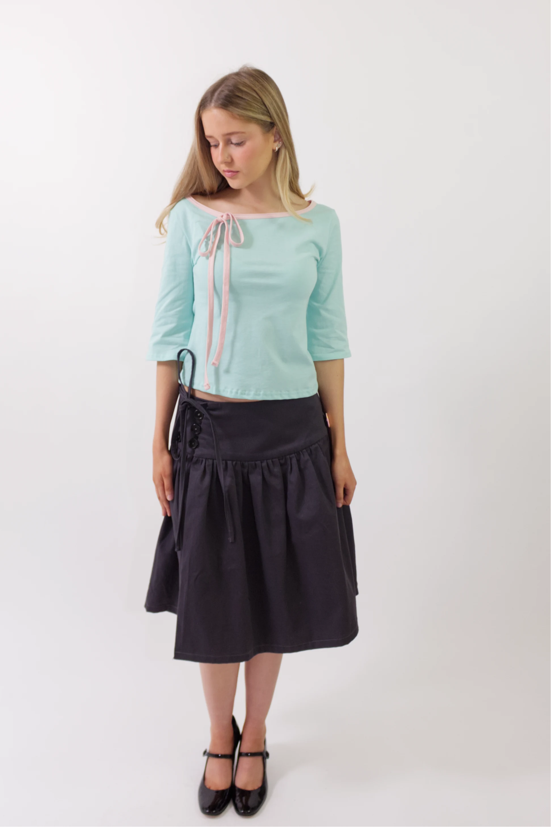 A woman modeling a light mint green three-quarter sleeve top with a pink and white striped tie detail at the neckline, paired with a dark navy midi skirt and black Mary Jane shoes, against a plain white studio background.