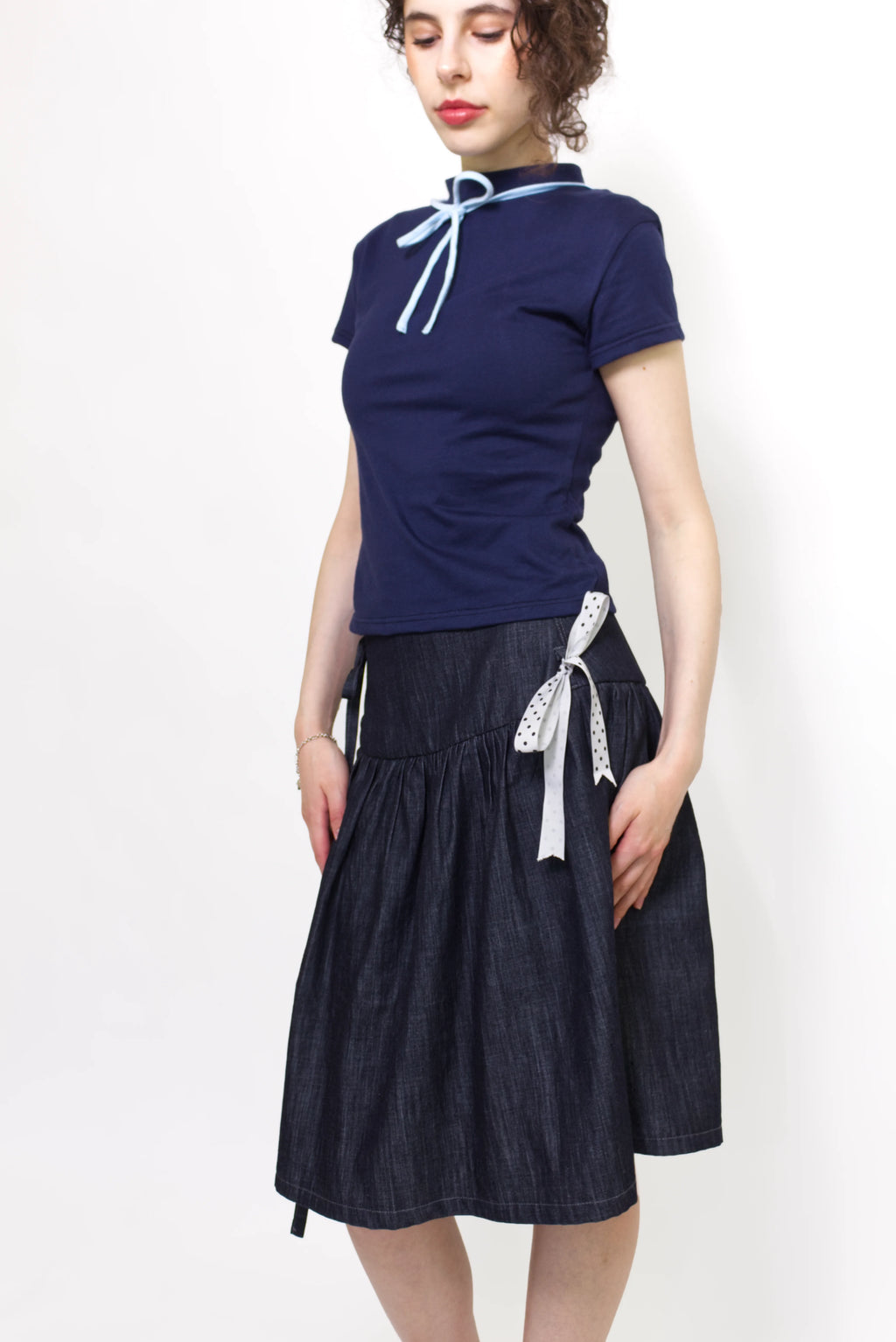 Woman wearing a navy and baby blue short sleeve top with a mock neck and blue bow with an indigo denim knee length gathered wrap skirt against a white background