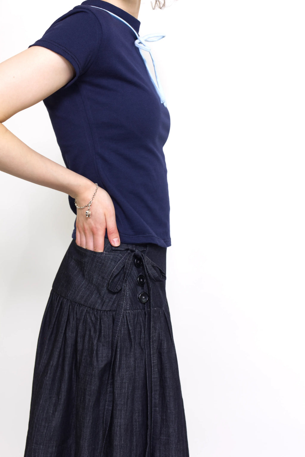 Woman wearing a navy and baby blue short sleeve top with a mock neck and blue bow with an indigo denim knee length gathered wrap skirt against a white background