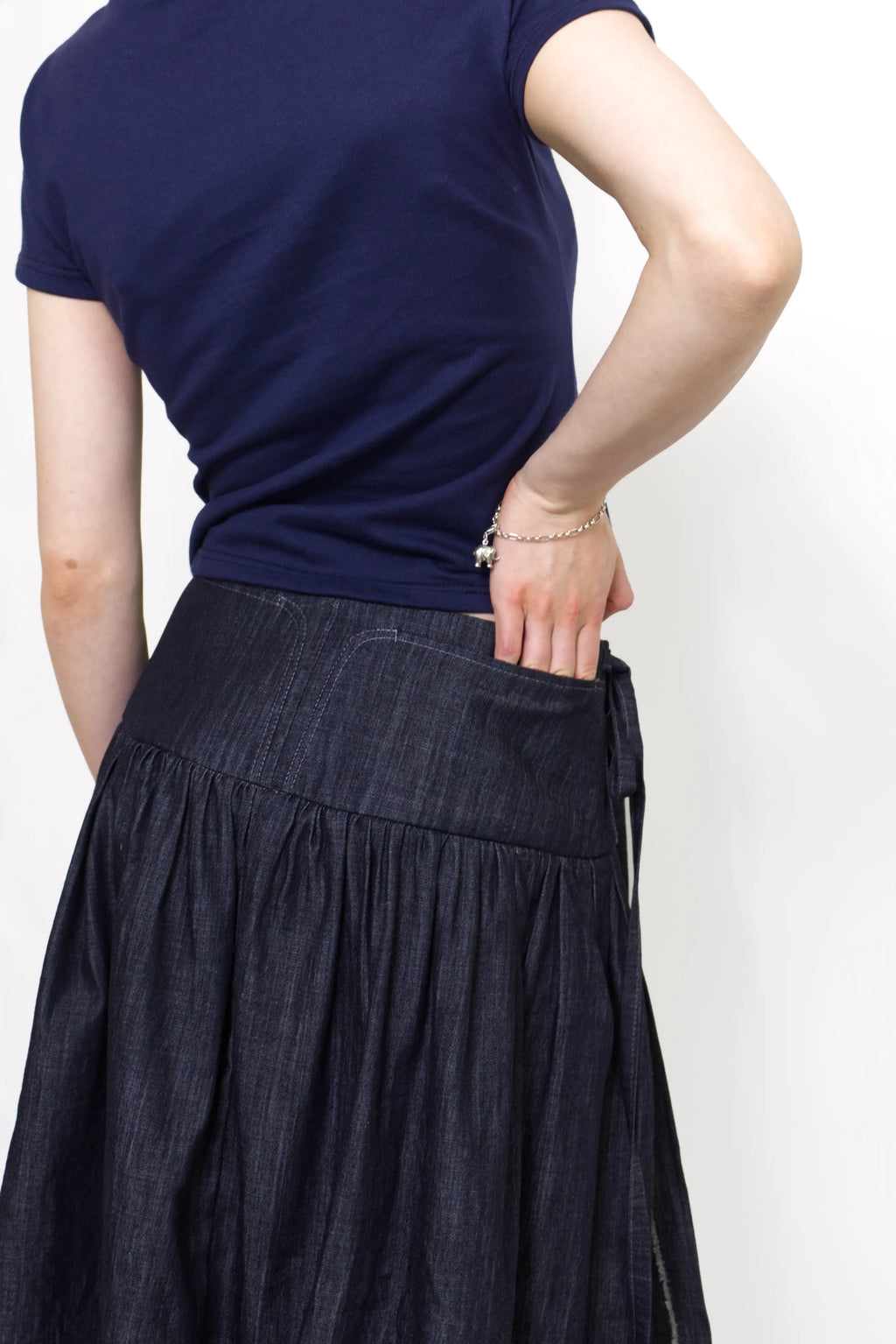 Woman wearing a navy and baby blue short sleeve top with a mock neck and blue bow with an indigo denim knee length gathered wrap skirt against a white background