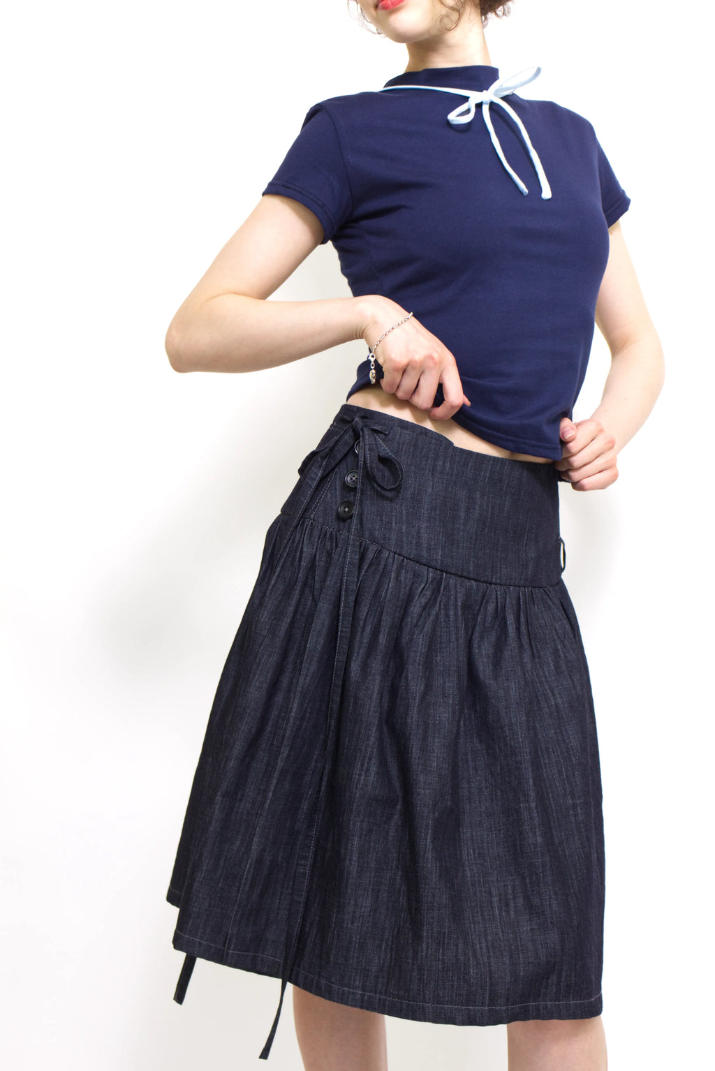 Woman wearing a navy and baby blue short sleeve top with a mock neck and blue bow with an indigo denim knee length gathered wrap skirt against a white background