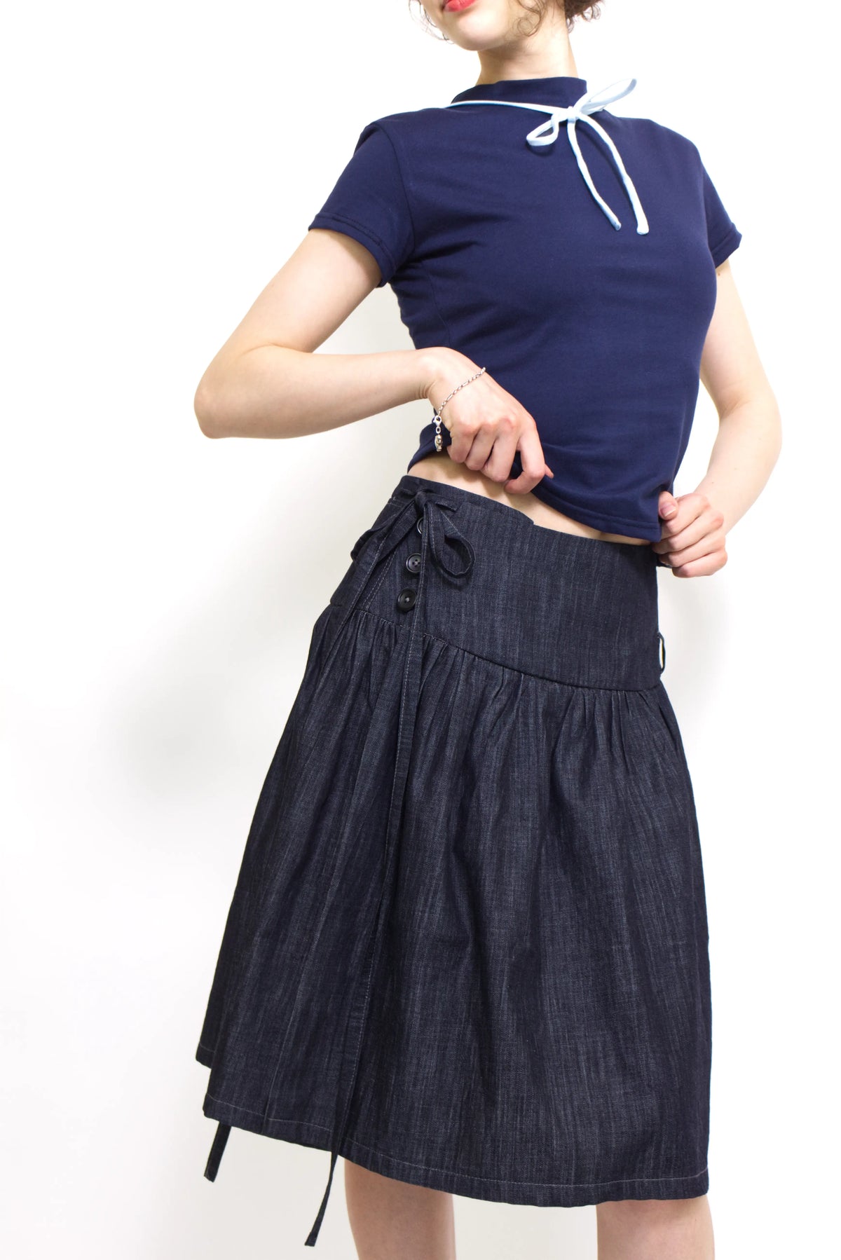 Woman wearing a navy and baby blue short sleeve top with a mock neck and blue bow with an indigo denim knee length gathered wrap skirt against a white background