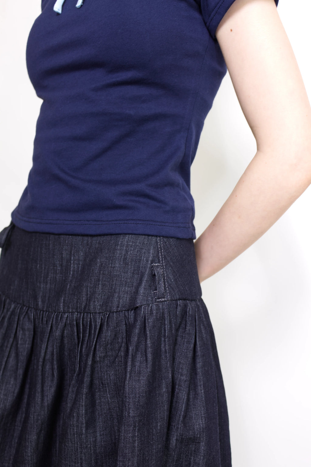 Woman wearing a navy and baby blue short sleeve top with a mock neck and blue bow with an indigo denim knee length gathered wrap skirt against a white background