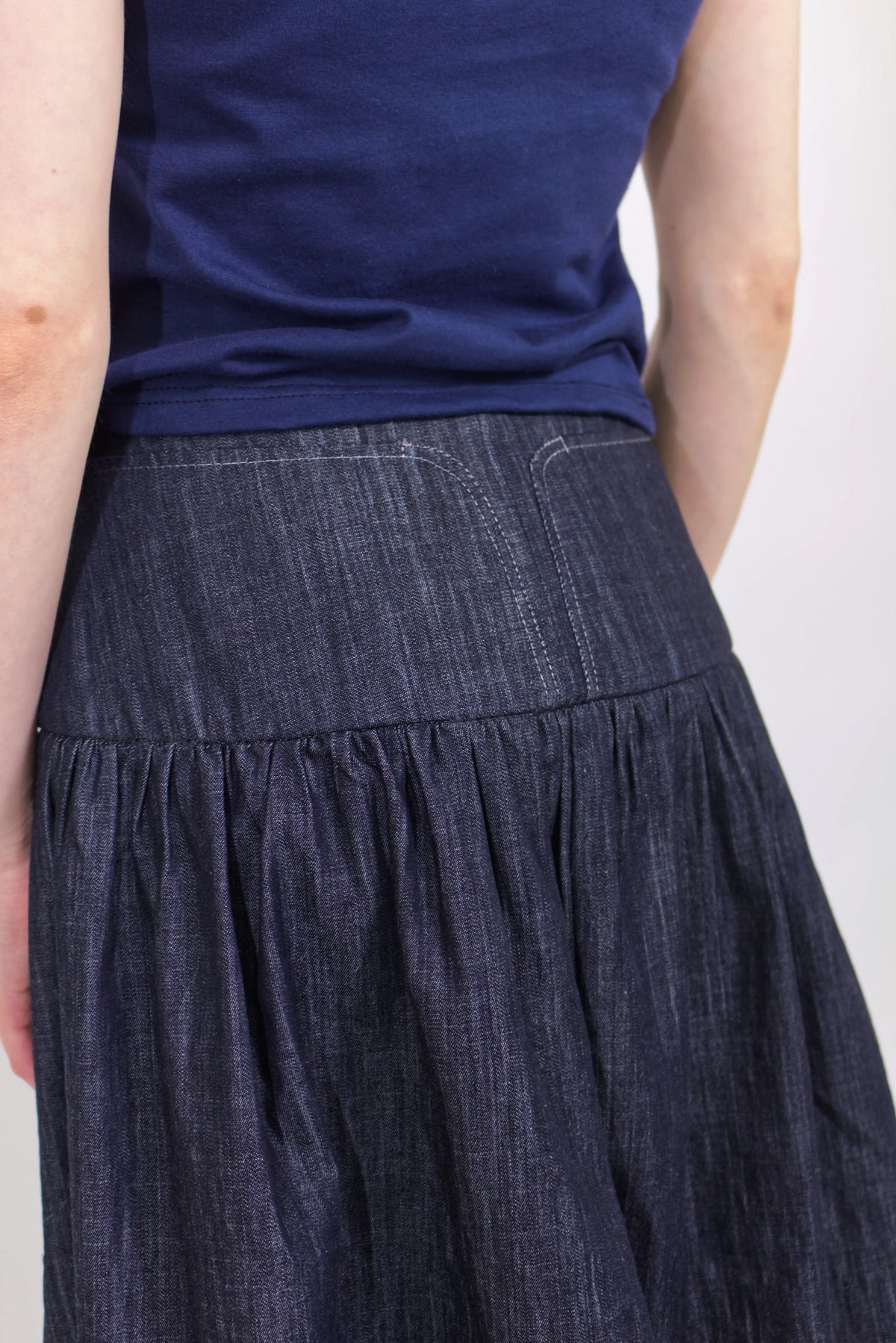 Woman wearing a navy and baby blue short sleeve top with a mock neck and blue bow with an indigo denim knee length gathered wrap skirt against a white background