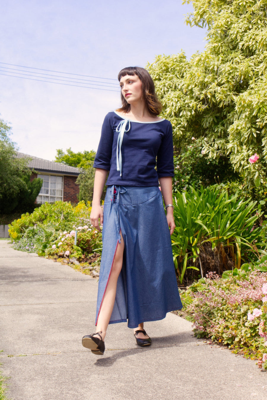 Woman in a blue  boat neck top and maxi wrap skirt outfit walking on a sidewalk with greenery in the background
