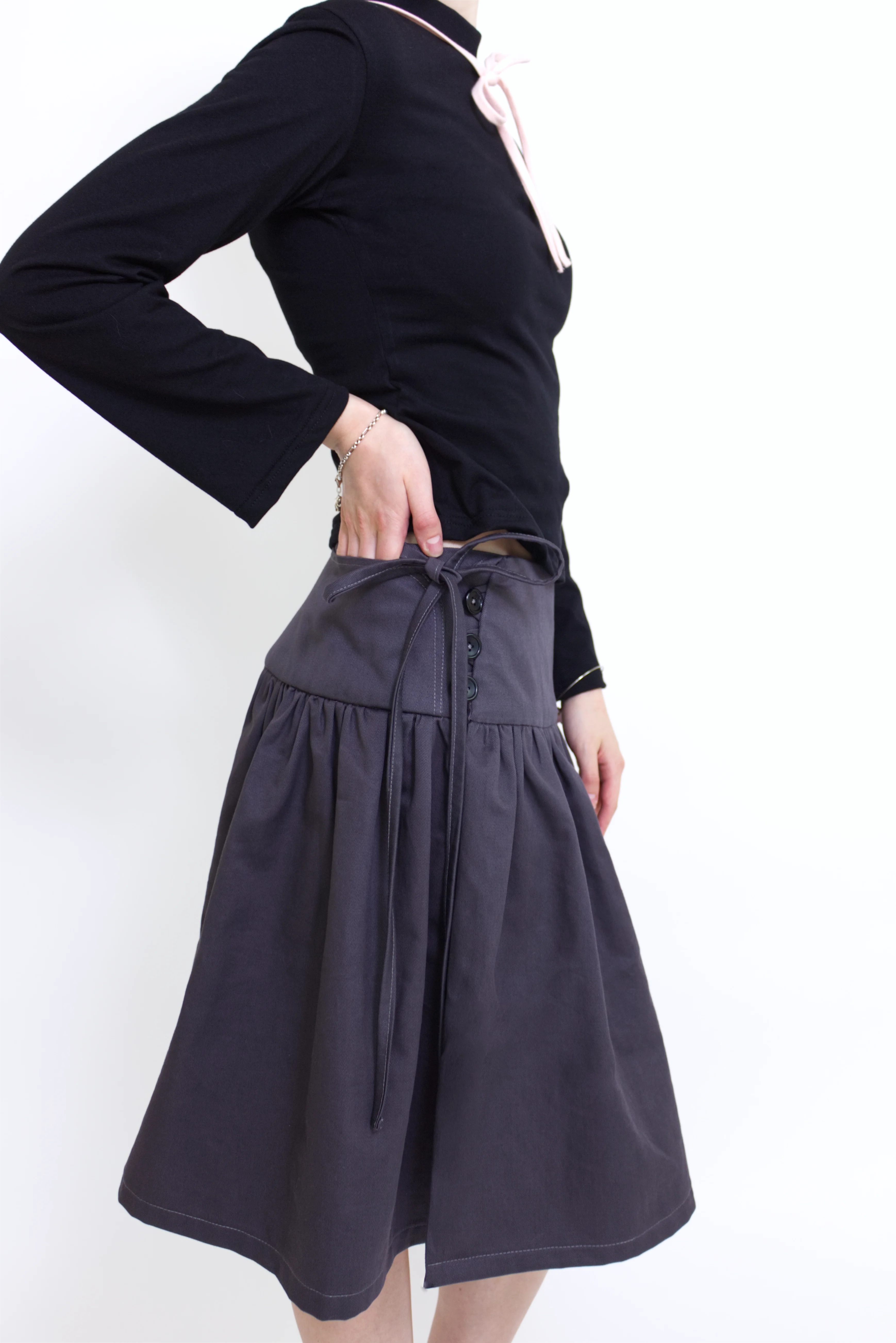 Woman wearing a black long sleeve top with a mock neck and pink bow with a  grey knee length gathered wrap skirt against a white background