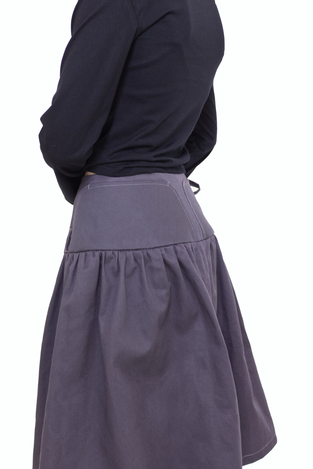 Woman wearing a black long sleeve top with a mock neck and pink bow with a  grey knee length gathered wrap skirt against a white background