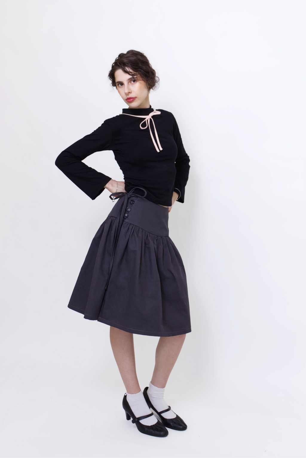 Woman wearing a black long sleeve top with a mock neck and pink bow with a  grey knee length gathered wrap skirt against a white background