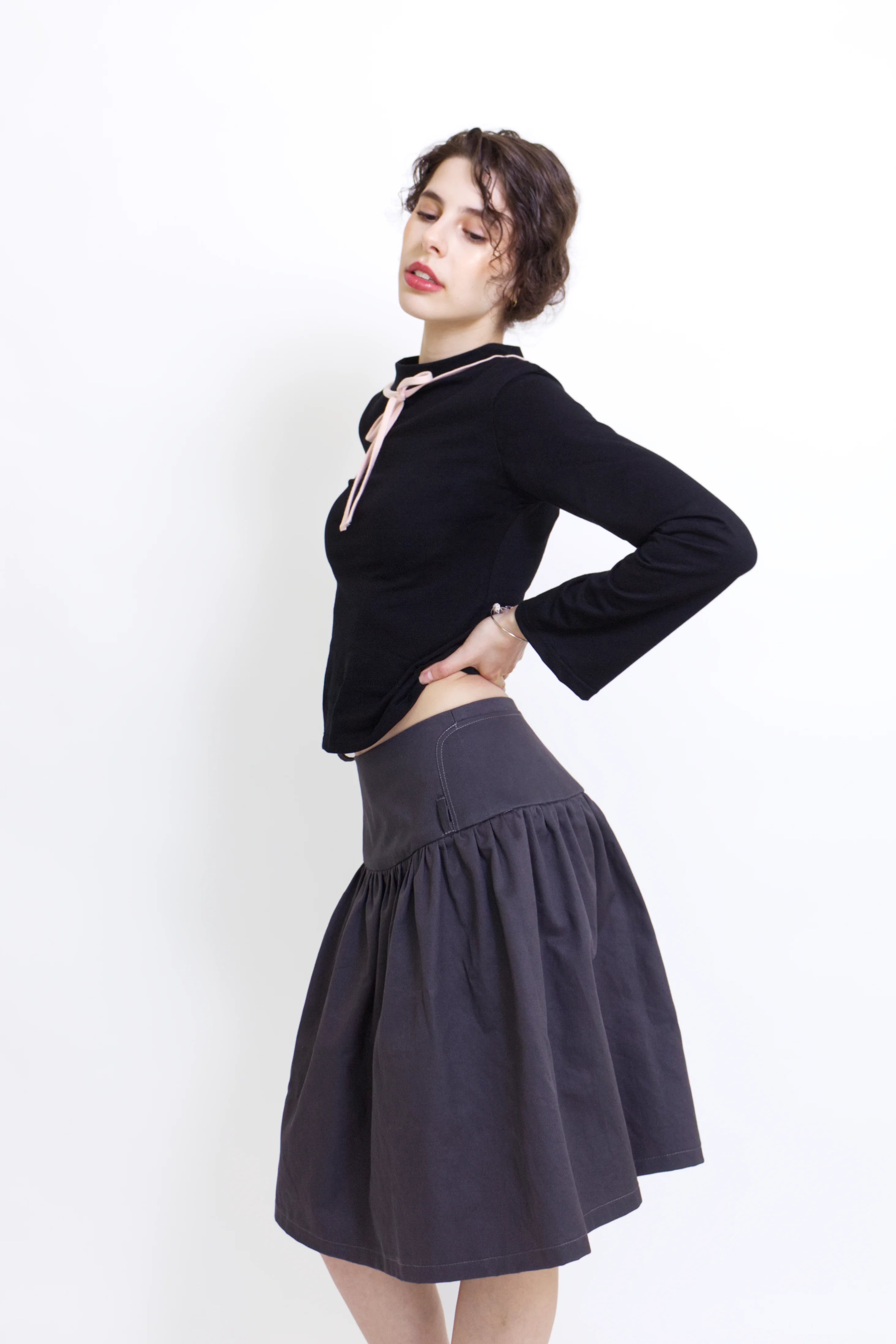 Woman wearing a black long sleeve top with a mock neck and pink bow with a  grey knee length gathered wrap skirt against a white background