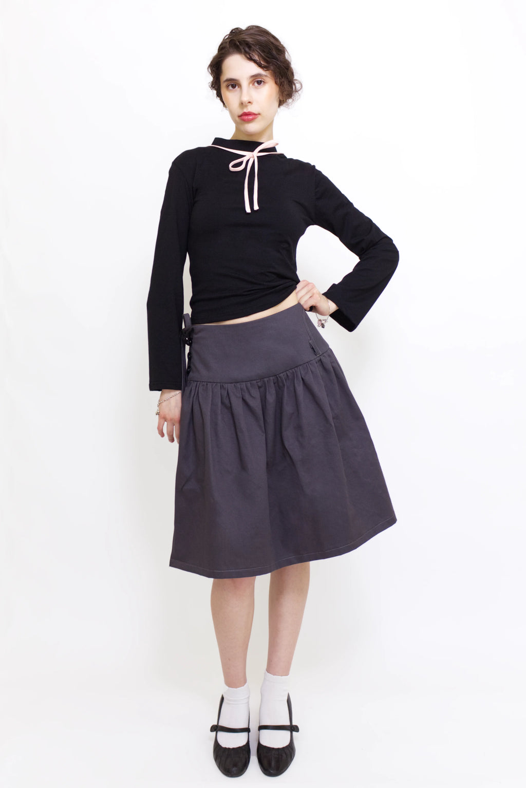 Woman wearing a black long sleeve top with a mock neck and pink bow with a  grey knee length gathered wrap skirt against a white background