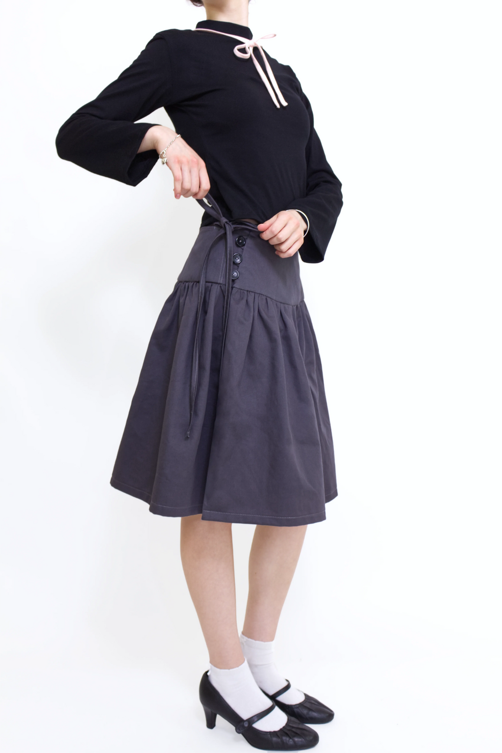 Woman wearing a black long sleeve top with a mock neck and pink bow with a  grey knee length gathered wrap skirt against a white background