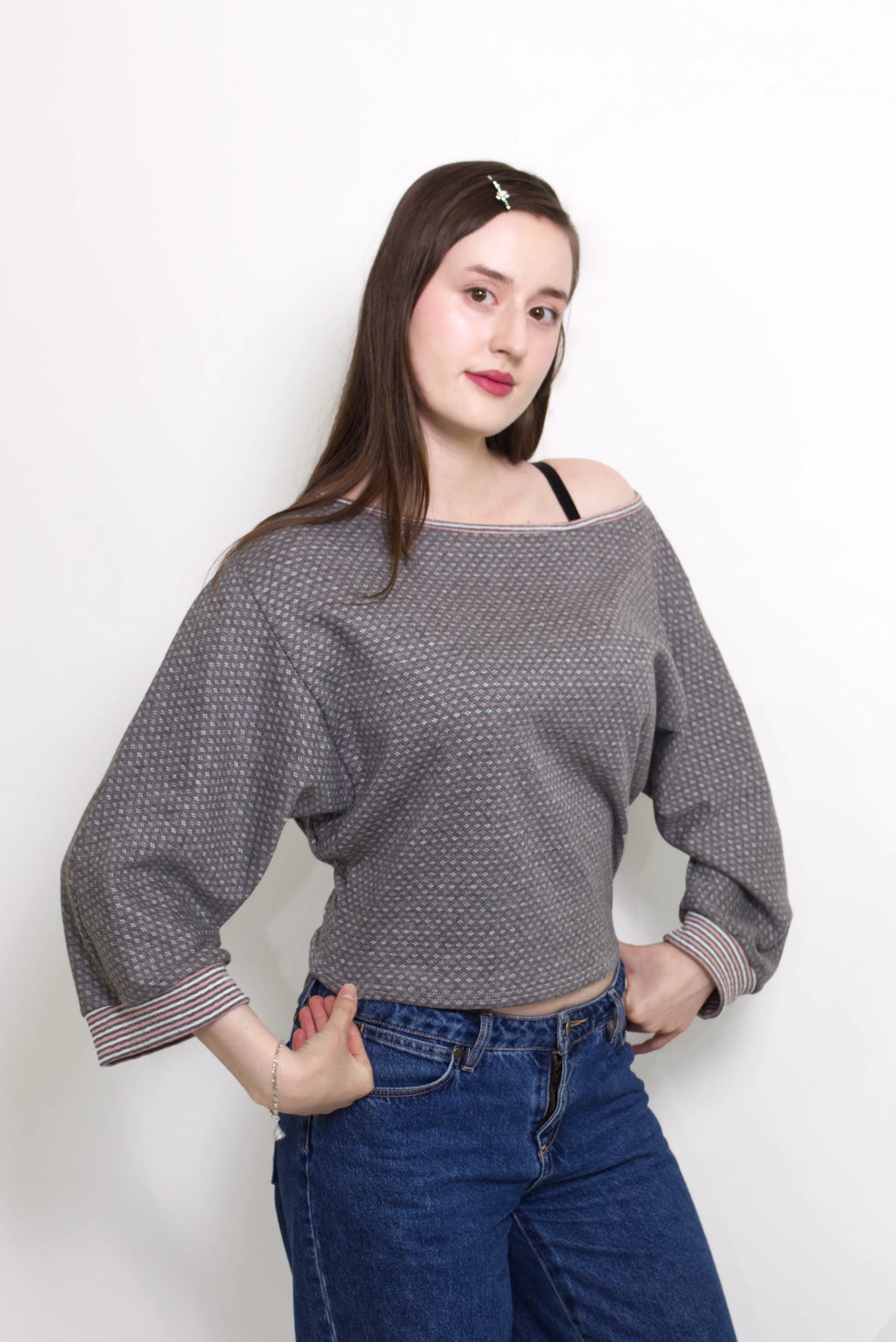 young woman wearing a gray, pink and white patterned oversized cotton boat neck pullover top, with long sleeves on a white background