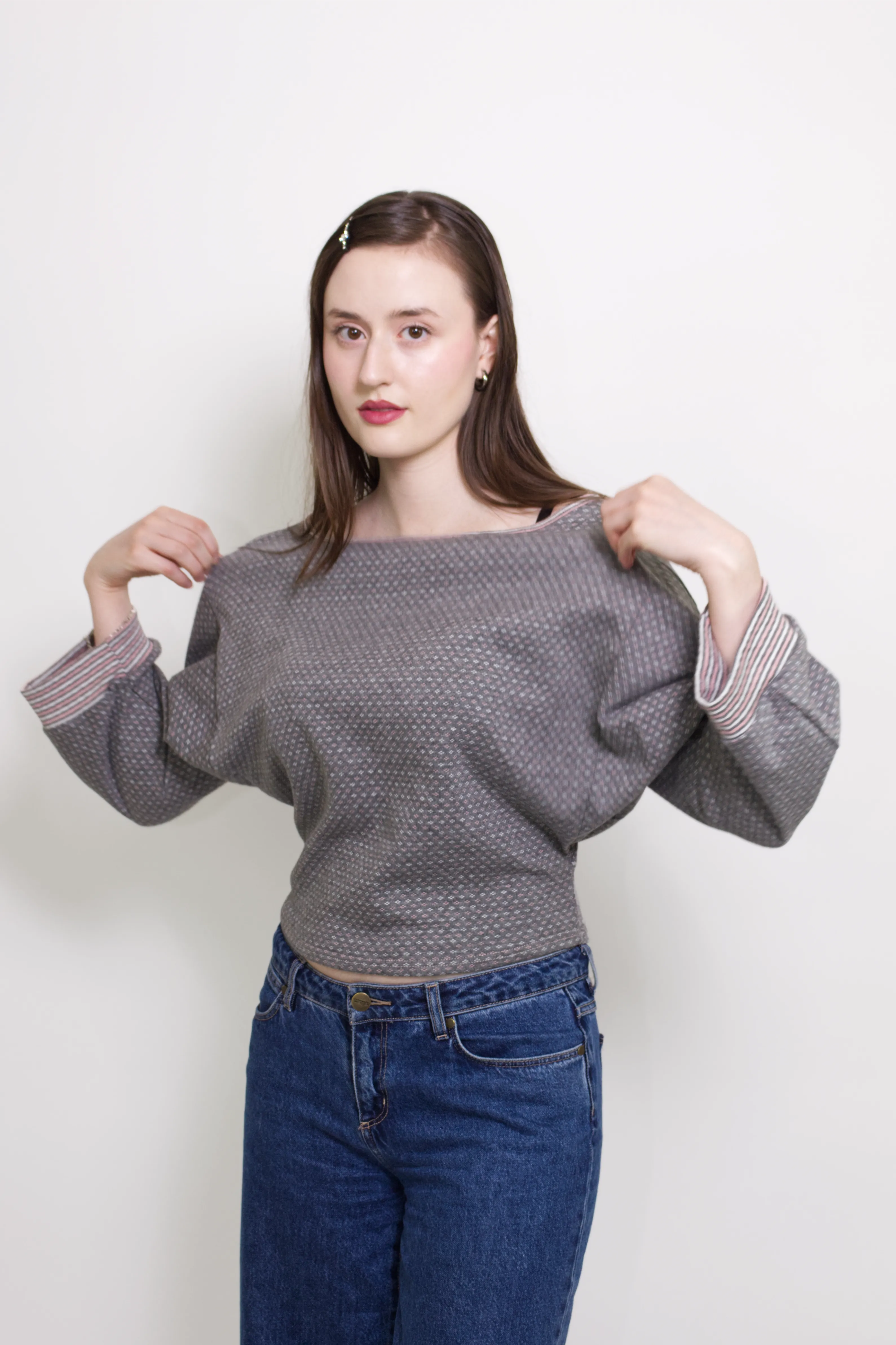 young woman wearing a grEy, pink and white patterned oversized cotton boat neck pullover top, with long sleeves on a white background