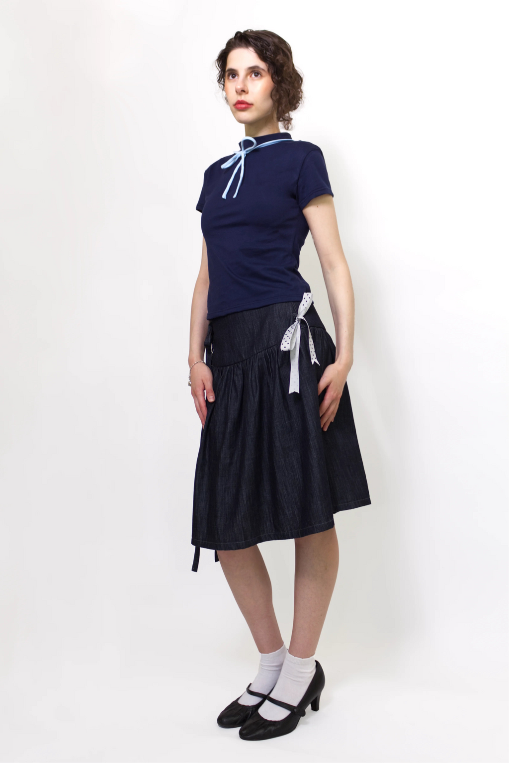 Woman wearing a navy and baby blue short sleeve top with a mock neck and blue bow with an indigo denim knee length gathered wrap skirt against a white background
