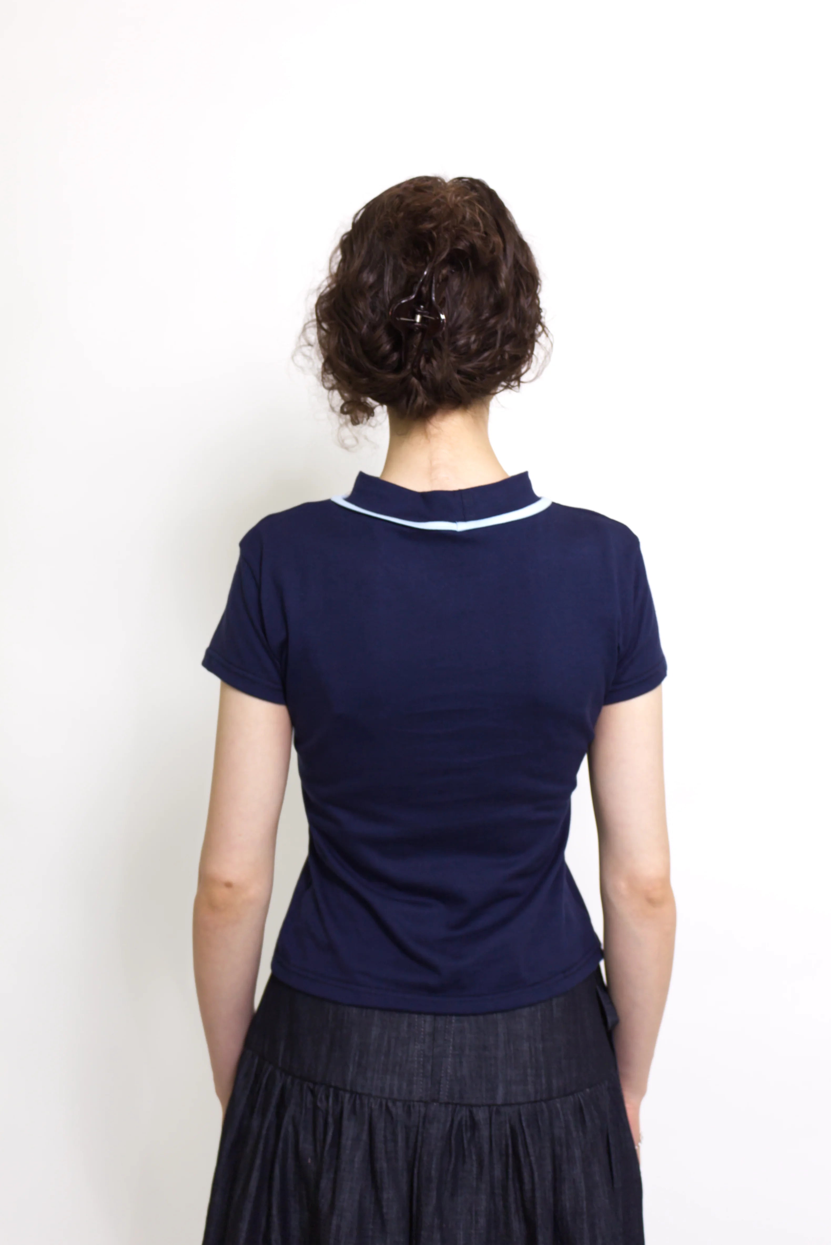 Woman wearing a navy and baby blue short sleeve top with a mock neck and blue bow with an indigo denim knee length gathered wrap skirt against a white background