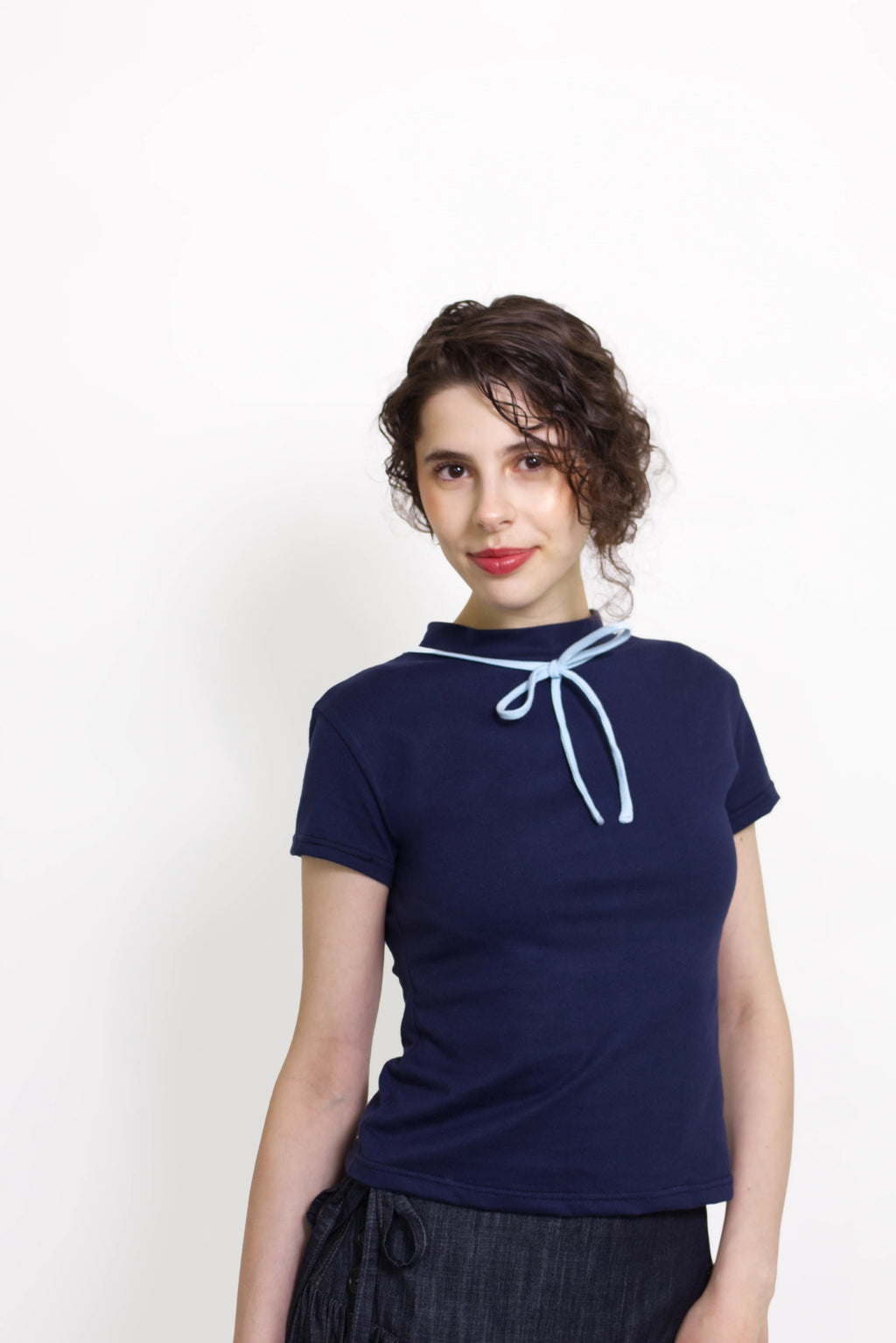 Woman wearing a navy and baby blue short sleeve top with a mock neck and blue bow with an indigo denim knee length gathered wrap skirt against a white background