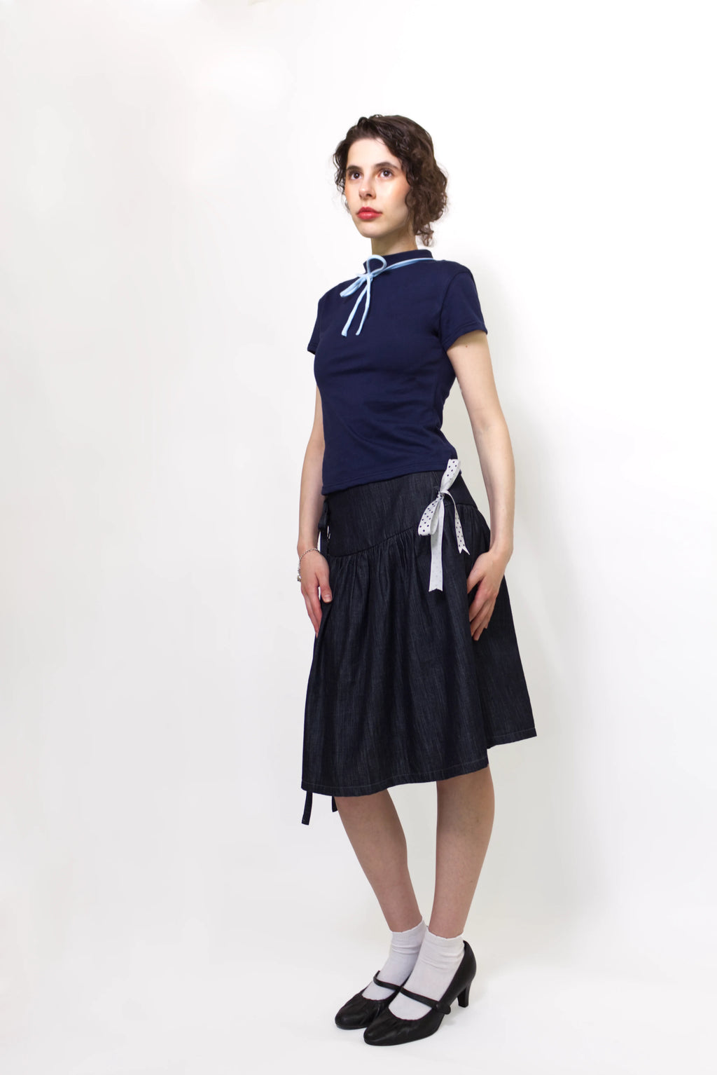 Woman wearing a navy and baby blue short sleeve top with a mock neck and blue bow with an indigo denim knee length gathered wrap skirt against a white background