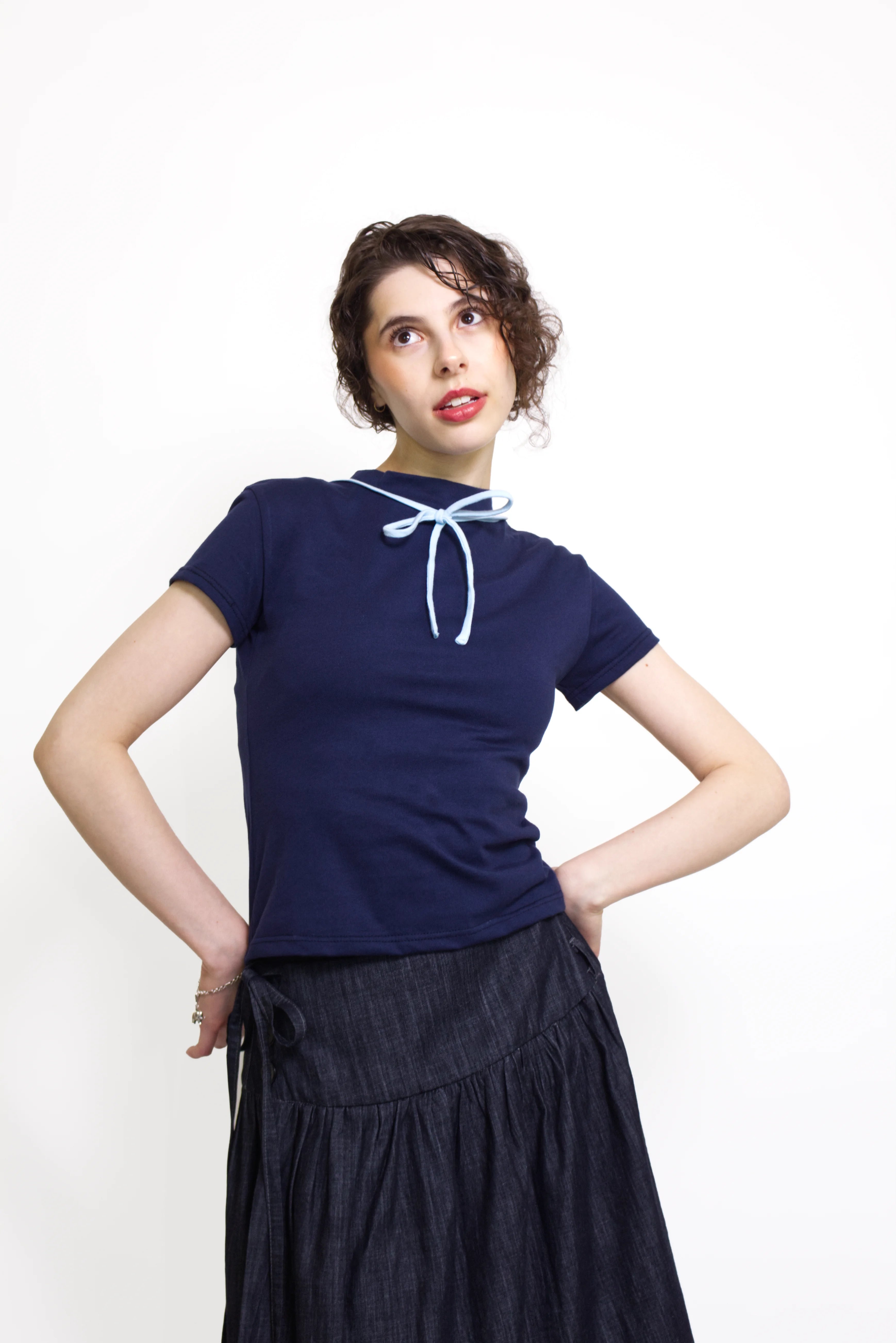 Woman wearing a navy and baby blue short sleeve top with a mock neck and blue bow with an indigo denim knee length gathered wrap skirt against a white background