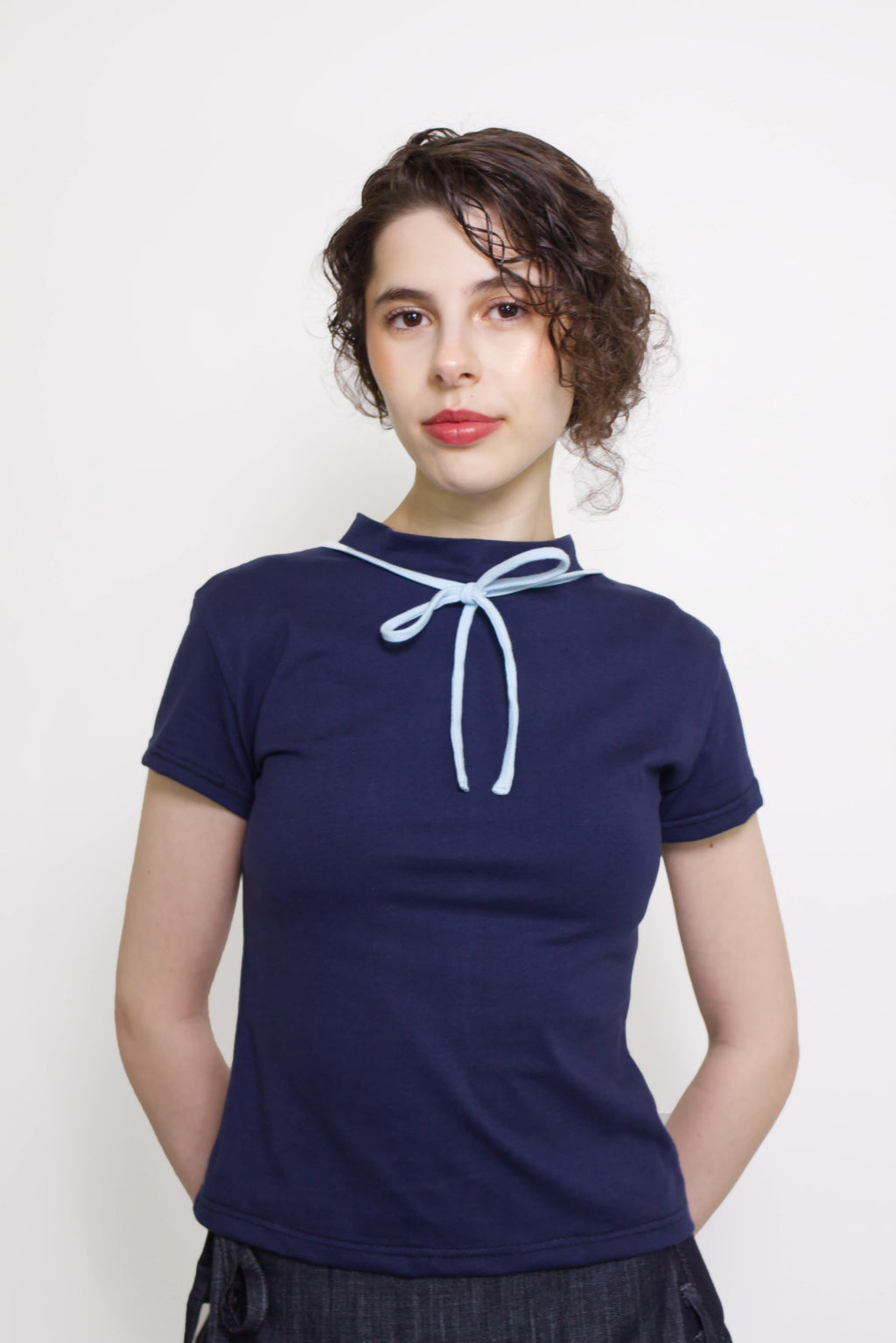 Woman wearing a navy and baby blue short sleeve top with a mock neck and blue bow with an indigo denim knee length gathered wrap skirt against a white background