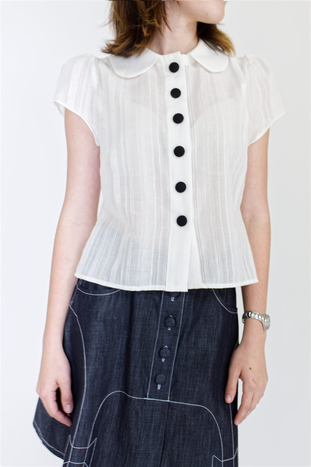 A young woman wearing a sheer ivory blouse with a peter pan collar, gathered cap sleeve and black buttons with a cute dark denim aline skirt with bow details and pockets.