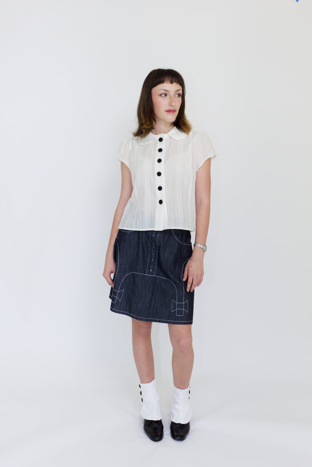 A young woman wearing a sheer ivory blouse with a peter pan collar, gathered cap sleeve and black buttons with a cute dark denim aline skirt with bow details and pockets.