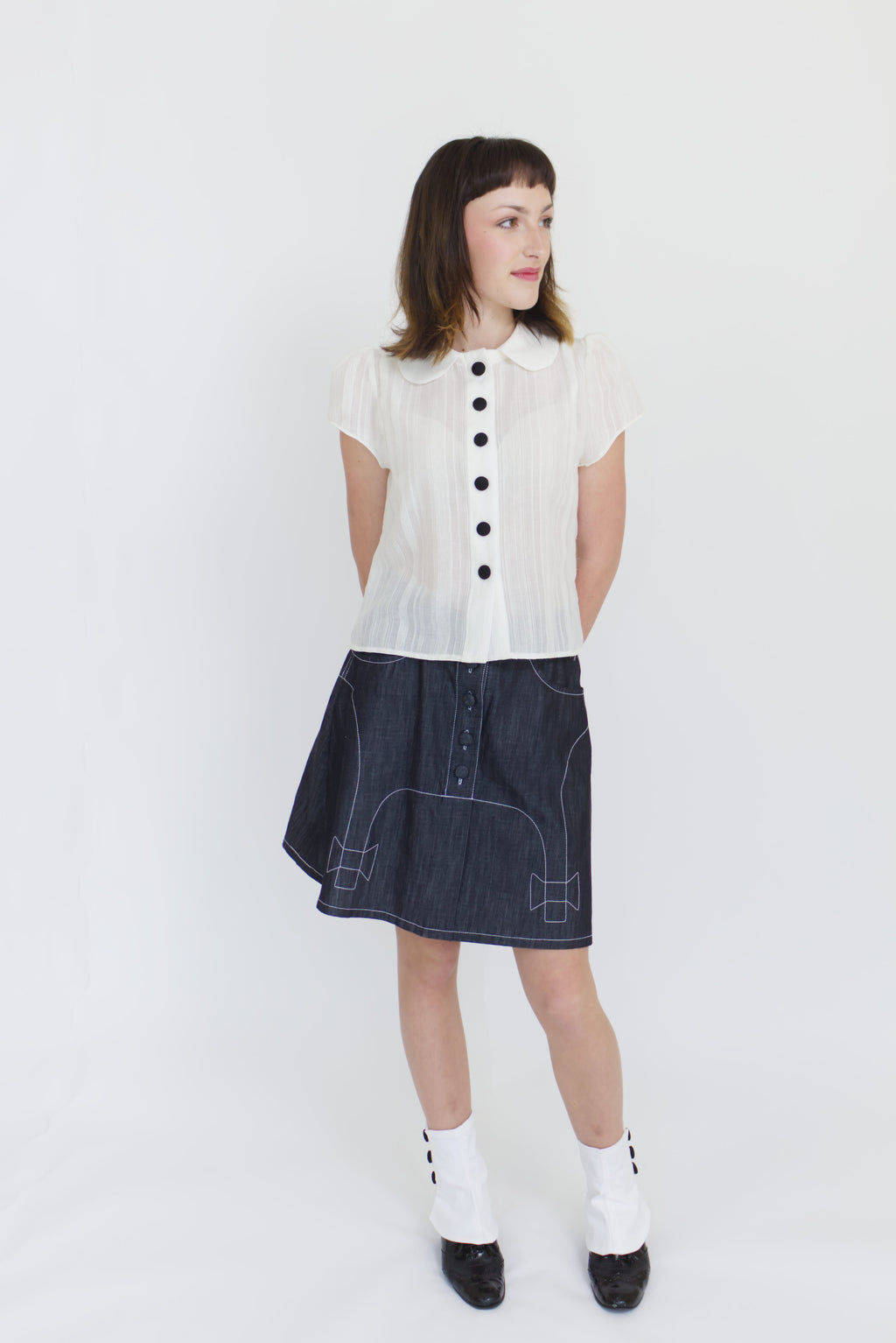 A young woman wearing a sheer ivory blouse with a peter pan collar, gathered cap sleeve and black buttons with a cute dark denim aline skirt with bow details and pockets.