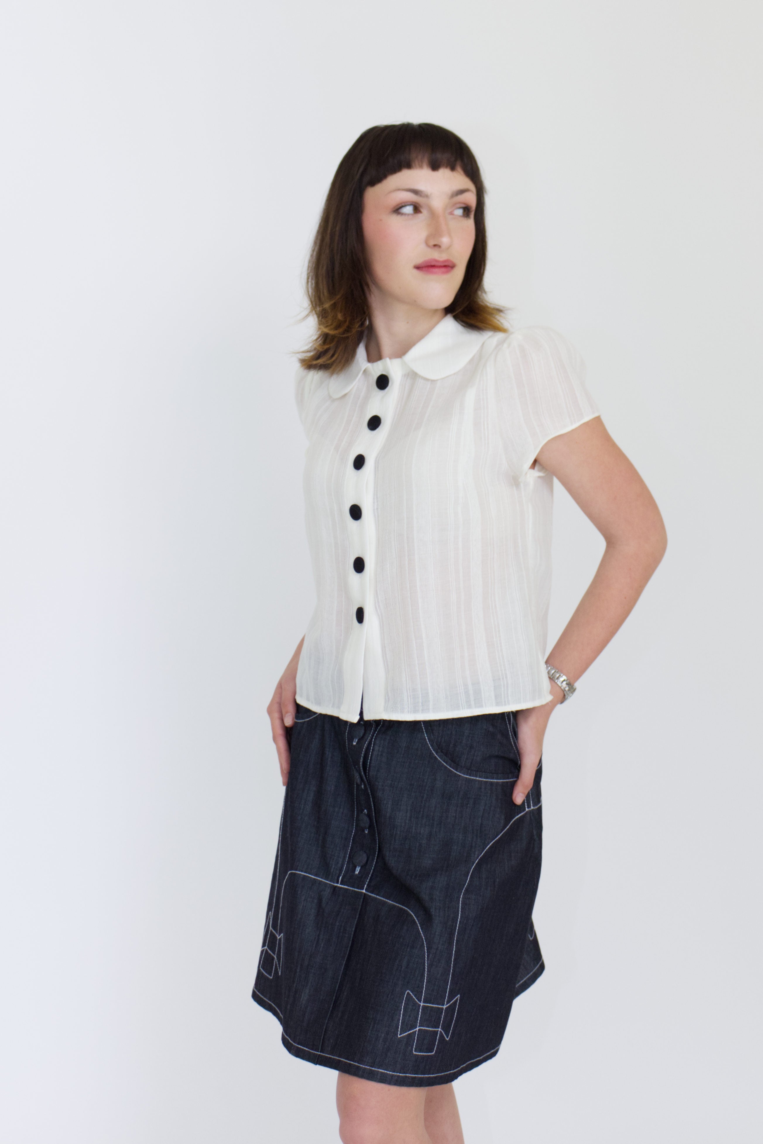 A young woman wearing a sheer ivory blouse with a peter pan collar, gathered cap sleeve and black buttons with a cute dark denim aline skirt with bow details and pockets.