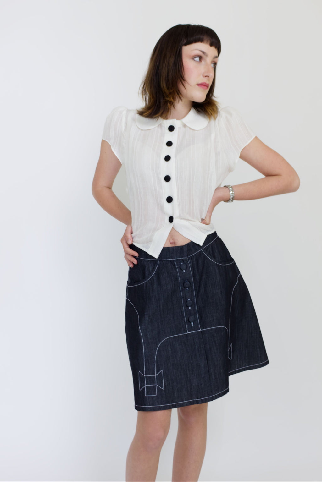 A young woman wearing a sheer ivory blouse with a peter pan collar, gathered cap sleeve and black buttons with a cute dark denim aline skirt with bow details and pockets.