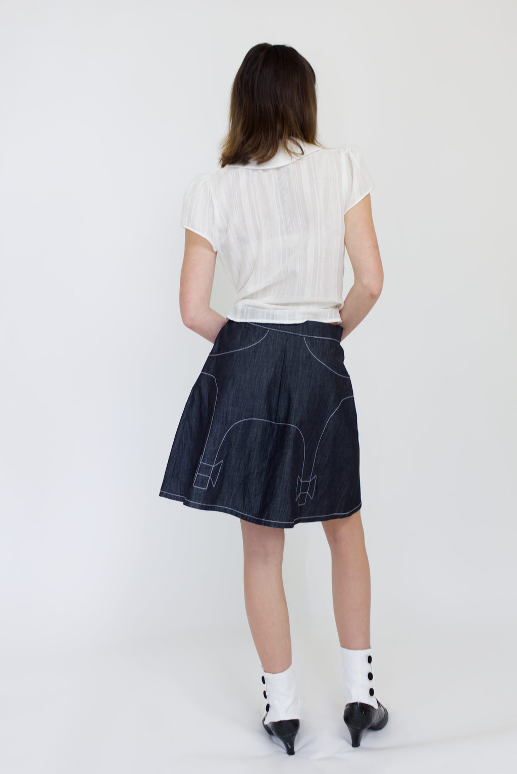 A young woman wearing a sheer ivory blouse with a peter pan collar, gathered cap sleeve and black buttons with a cute dark denim aline skirt with bow details and pockets.