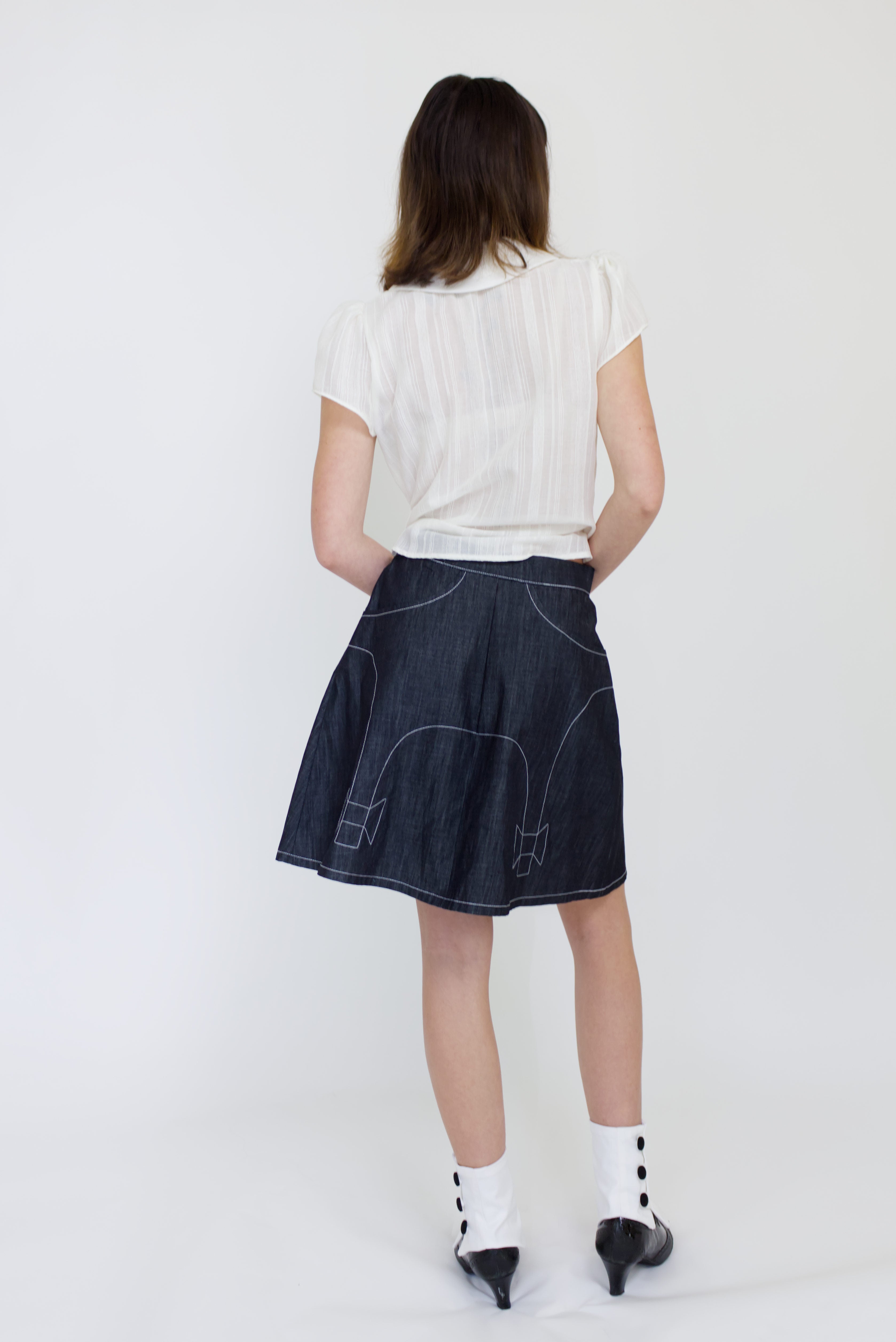 A young woman wearing a sheer ivory blouse with a peter pan collar, gathered cap sleeve and black buttons with a cute dark denim aline skirt with bow details and pockets.