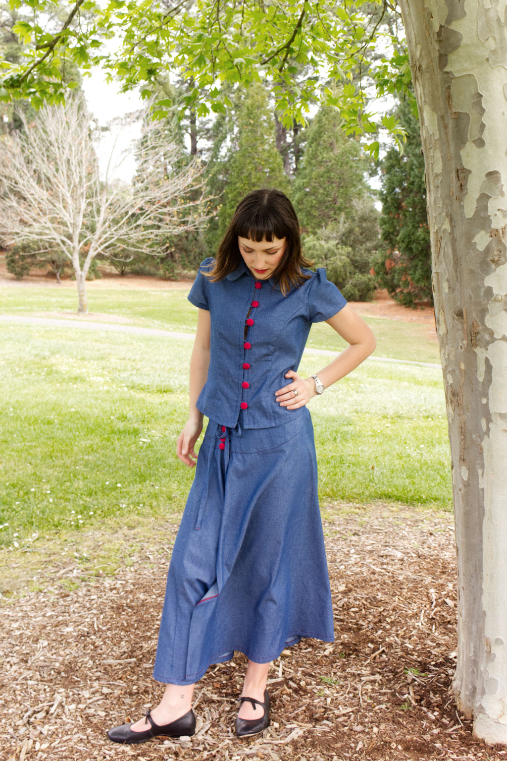 Young woman in a matching blue chambray cap sleeve princess line blouse and asymmetrical maxi warp skirt with a split, both with red buttons, walking in a park