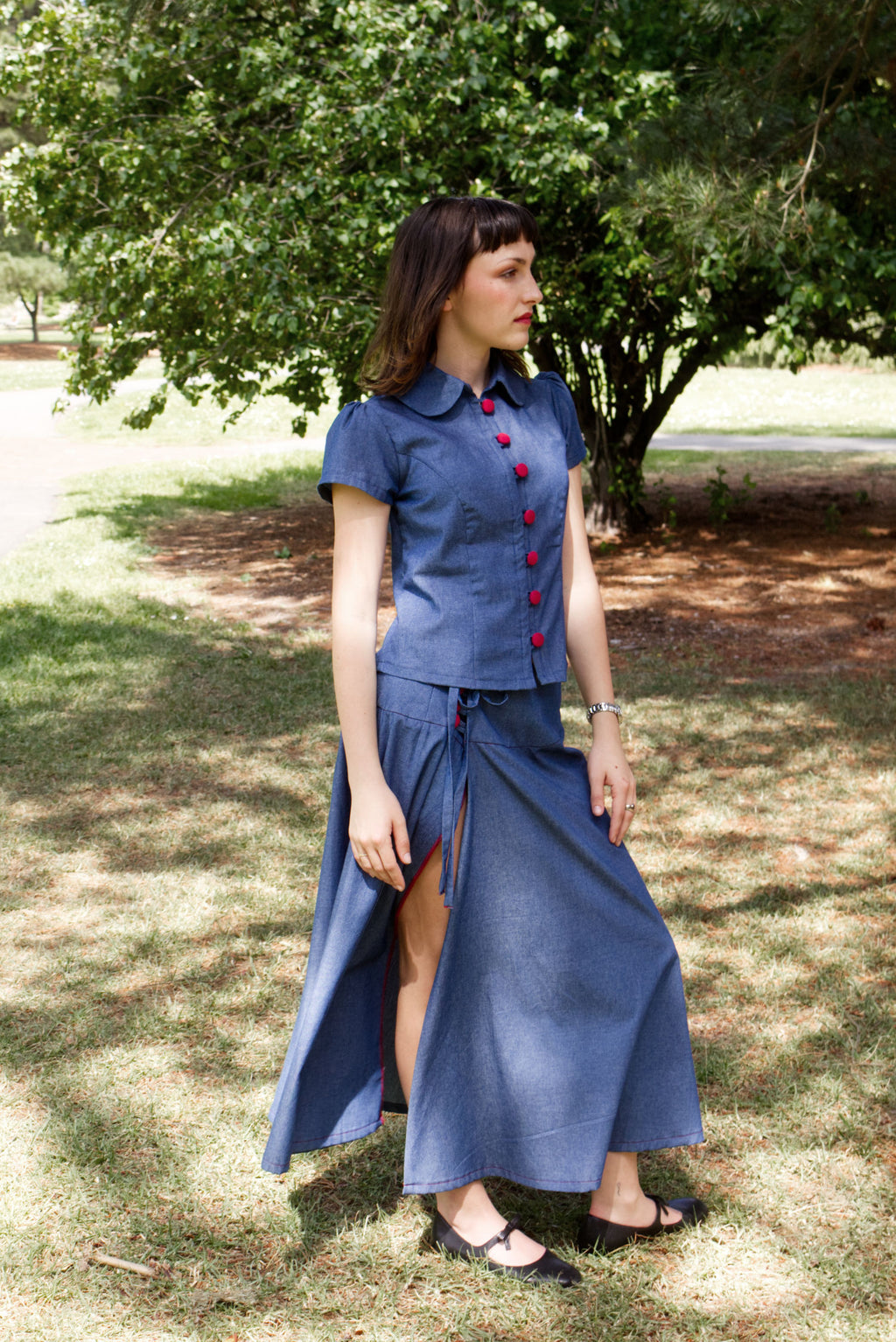 Young woman in a matching blue chambray cap sleeve princess line blouse and asymmetrical maxi warp skirt with a split, both with red buttons, walking in a park