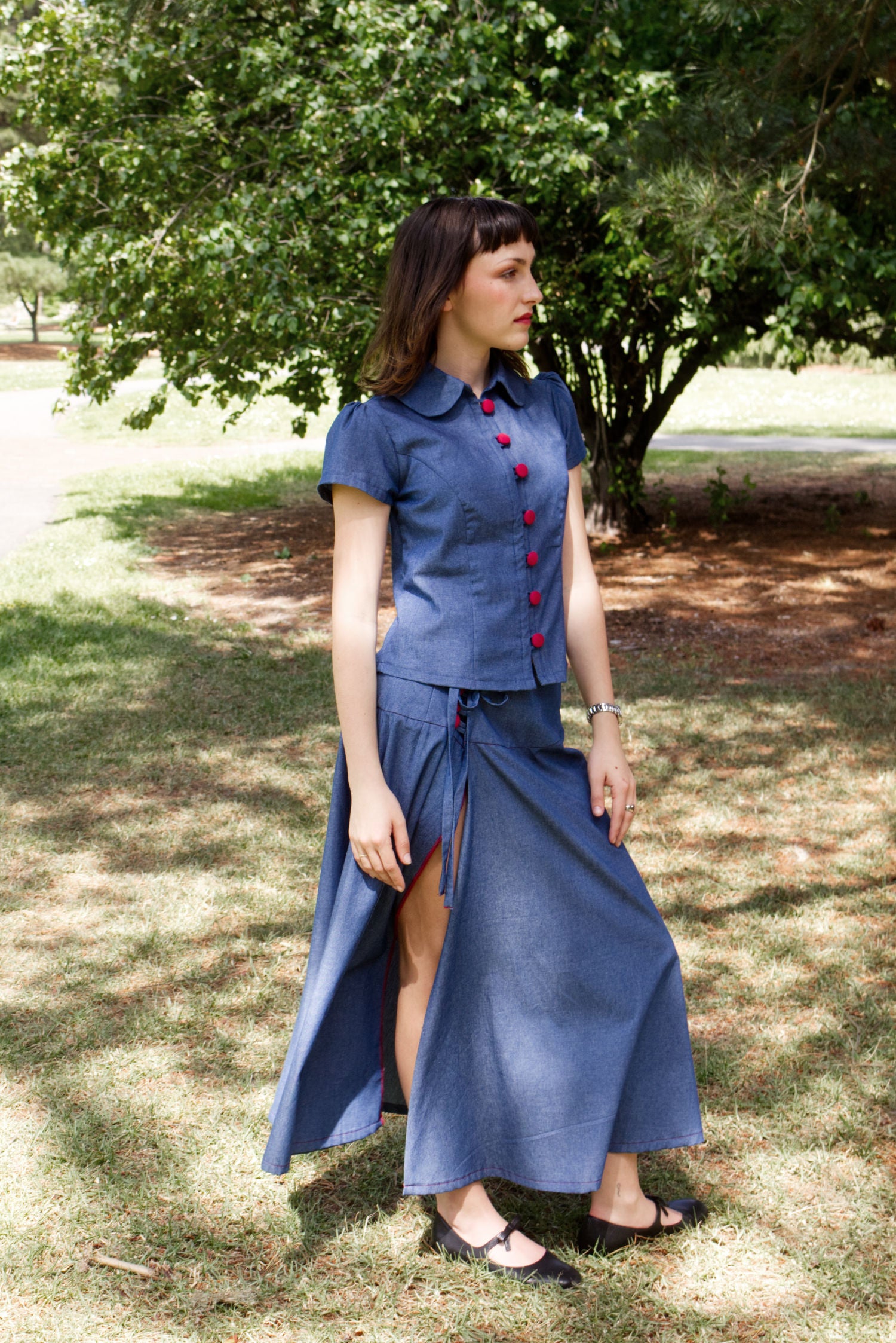 Young woman in a matching blue chambray cap sleeve princess line blouse and asymmetrical maxi warp skirt with a split, both with red buttons, walking in a park