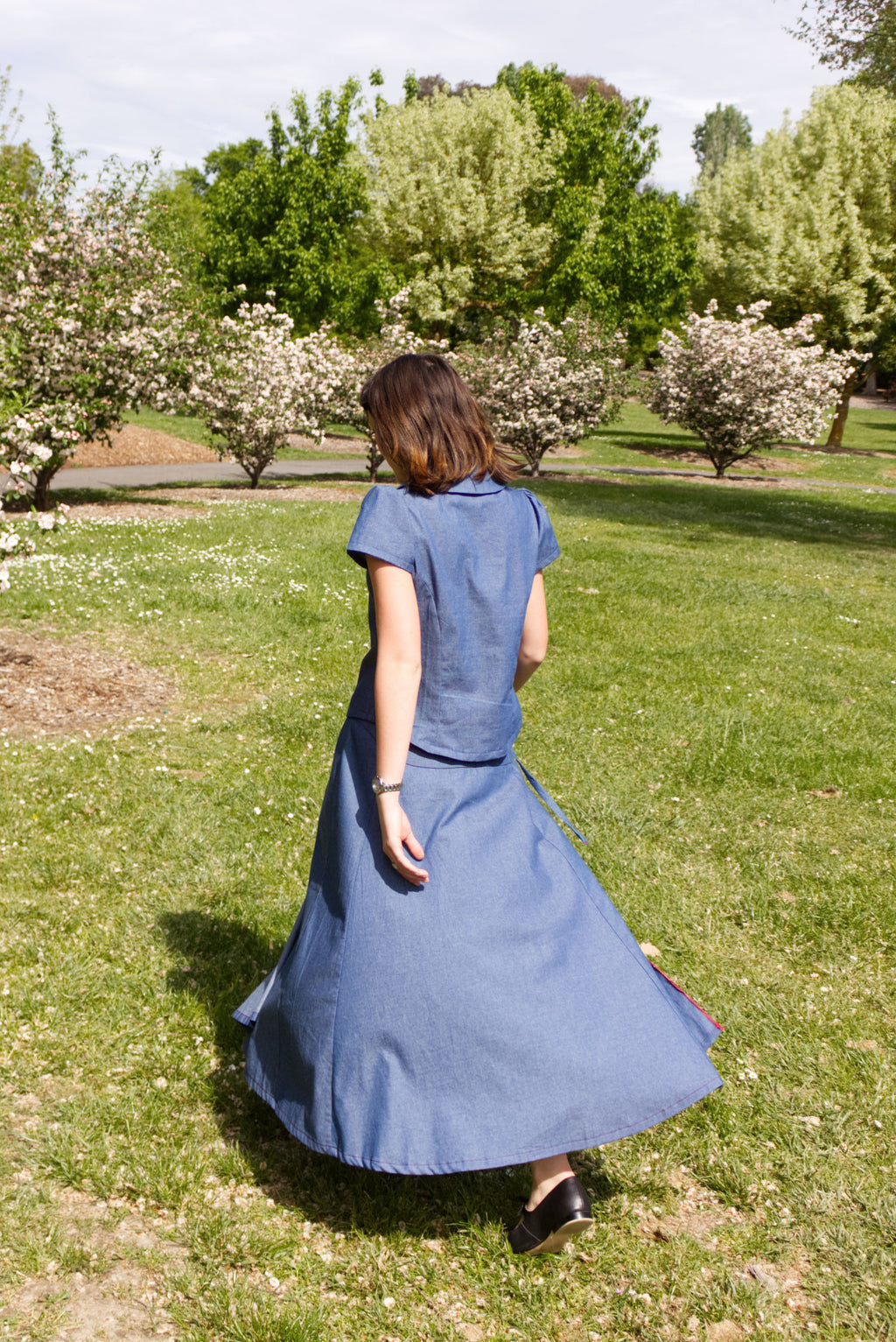 Young woman in a matching blue chambray cap sleeve princess line blouse and asymmetrical maxi warp skirt with a split, both with red buttons, walking in a park