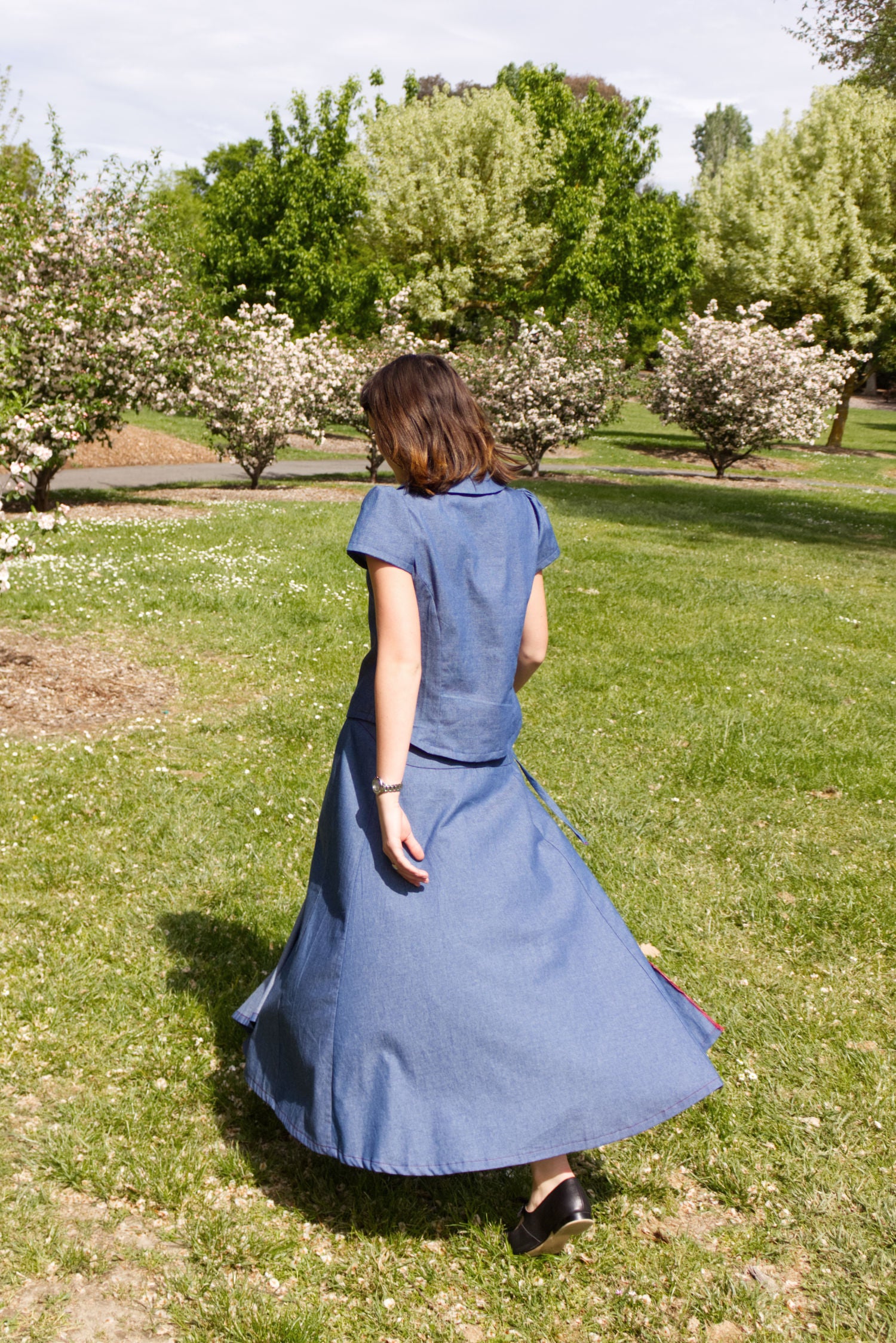 Young woman in a matching blue chambray cap sleeve princess line blouse and asymmetrical maxi warp skirt with a split, both with red buttons, walking in a park