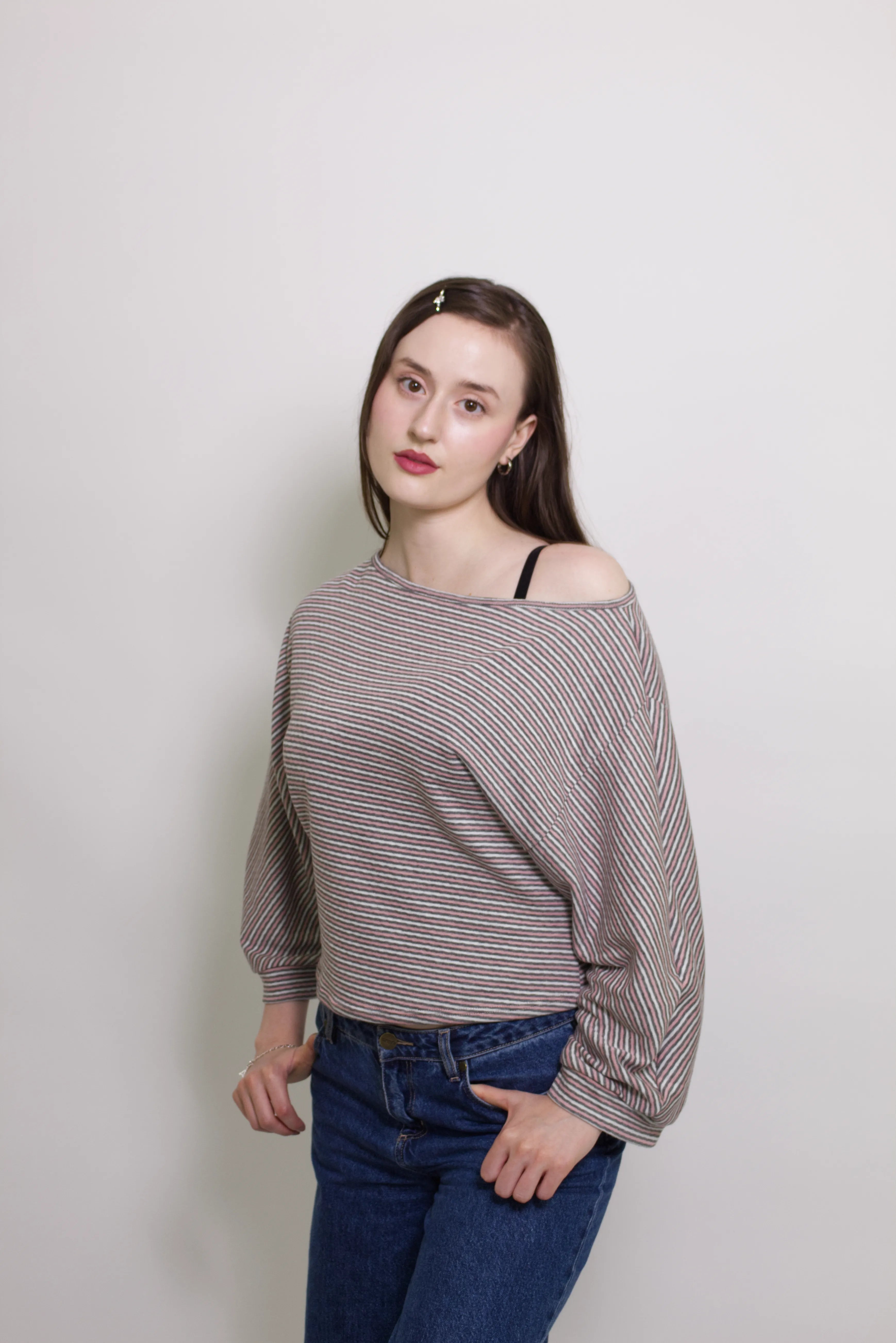 young woman wearing a pink striped oversized cotton boat neck pullover top, with long sleeves on a white background