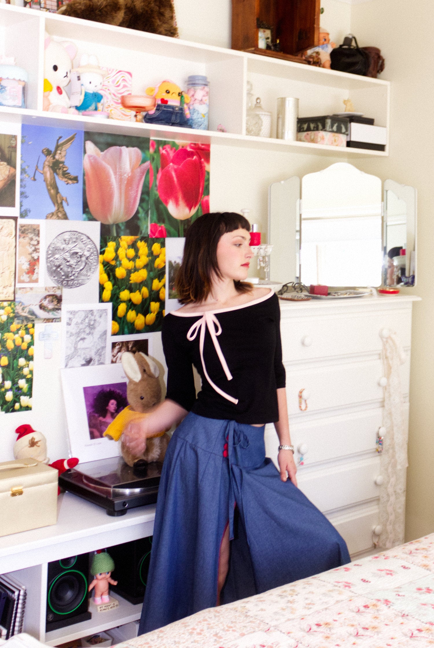 A young woman wearing a black and baby pink boat neck blouse with a ¾ sleeve, paired with a denim blue chambray maxi skirt, in a youthful, colourful bedroom listening to a Kate Bush vinyl record
