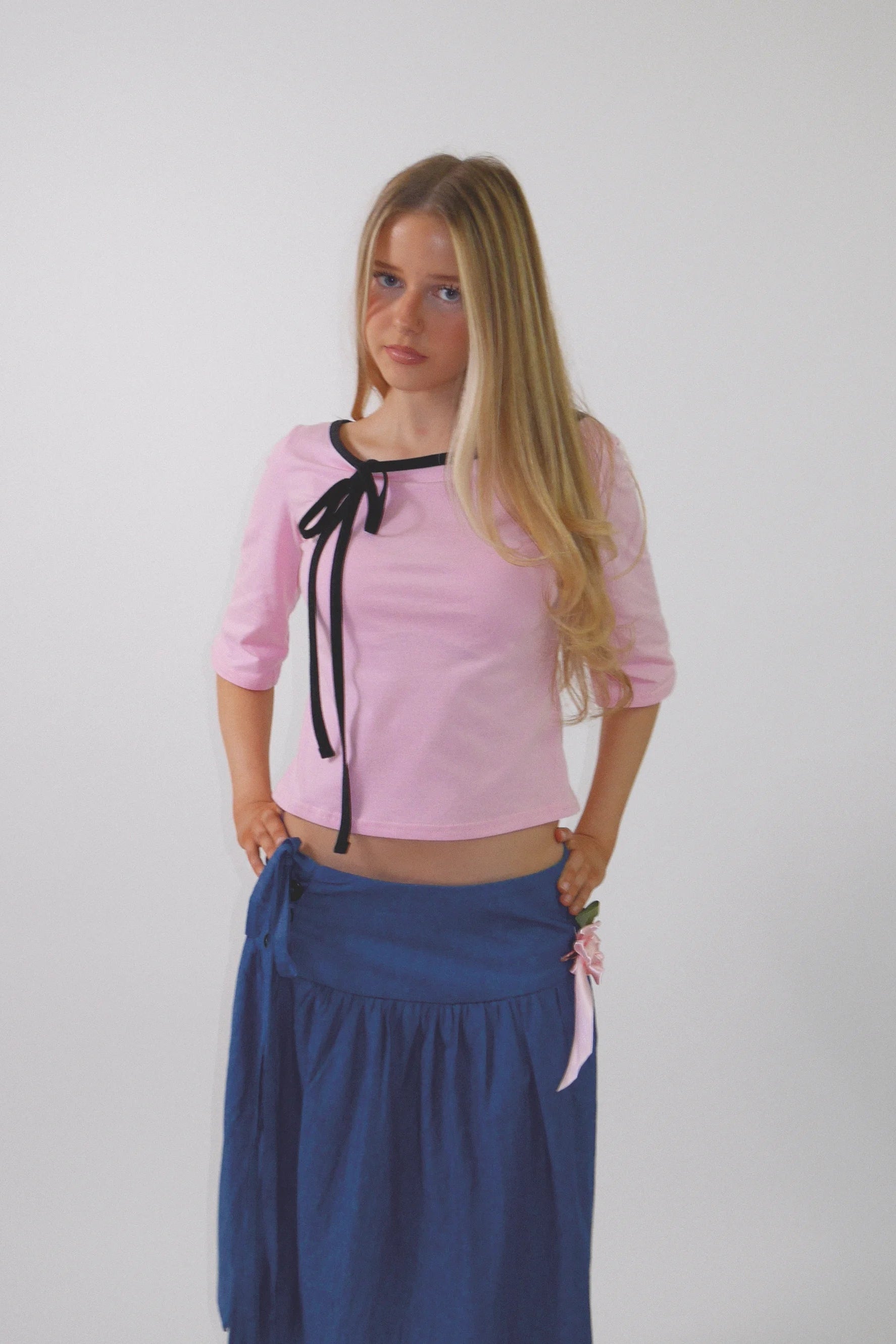 A young girl with long blonde hair stands against a light gray background wearing a light pink cropped top with black ribbon ties and a dark blue skirt.