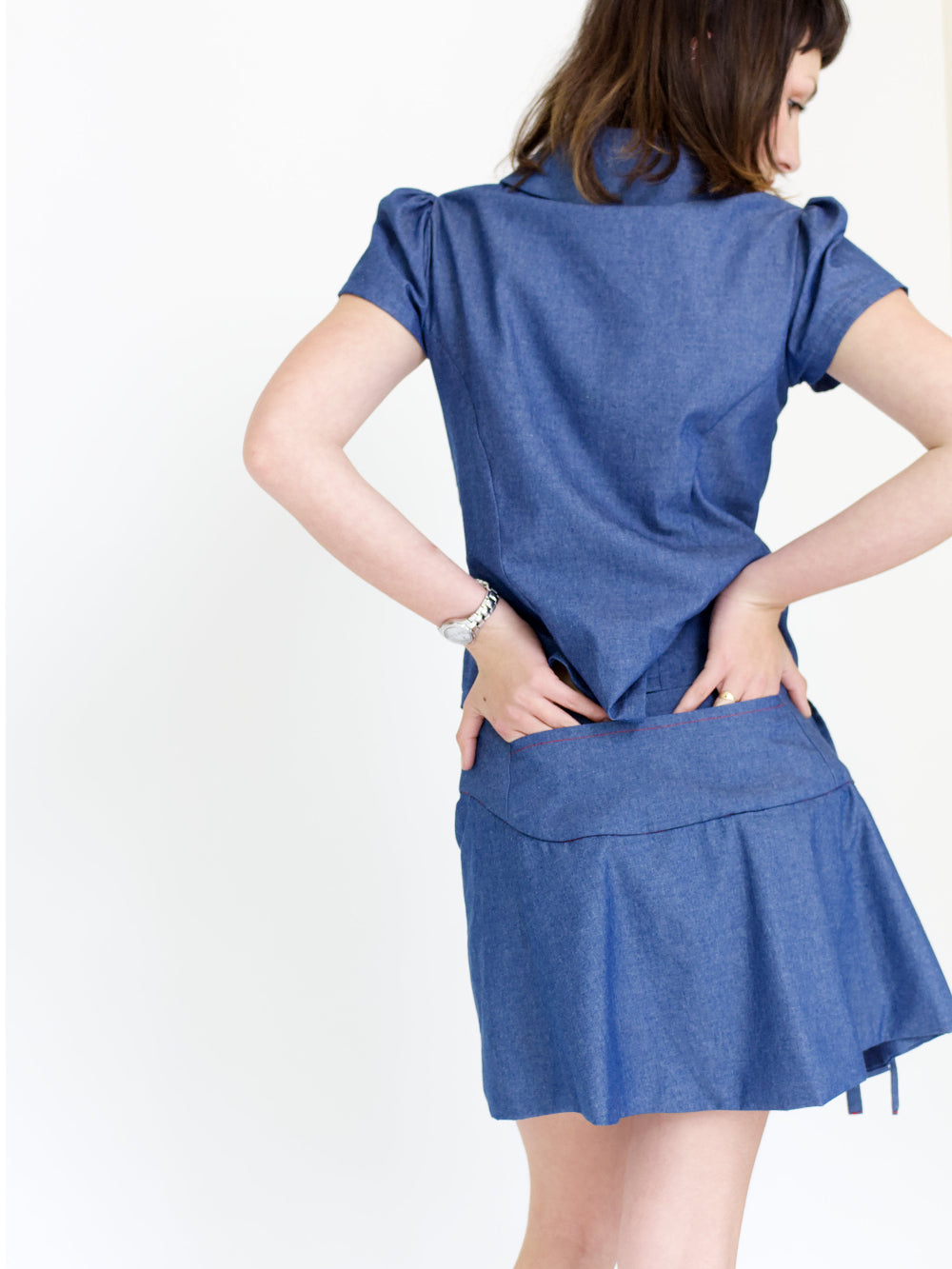 Young woman in a matching blue chambray cap sleeve, peter pan collar, princess line blouse and asymmetrical, low rise mini warp skirt, both with red buttons, in front of a white back ground