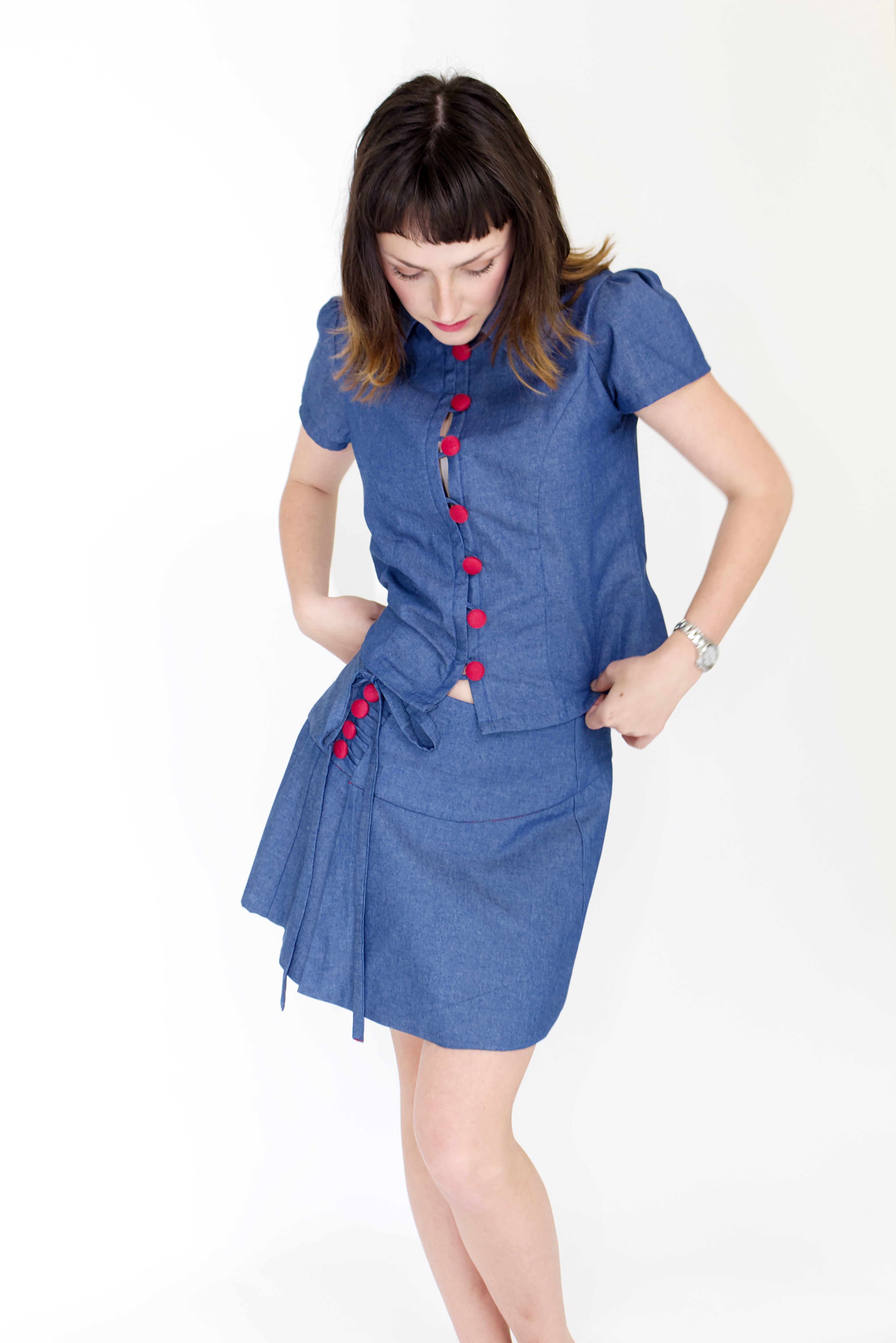 Young woman in a matching blue chambray cap sleeve, peter pan collar, princess line blouse and asymmetrical, low rise mini warp skirt, both with red buttons, in front of a white back ground