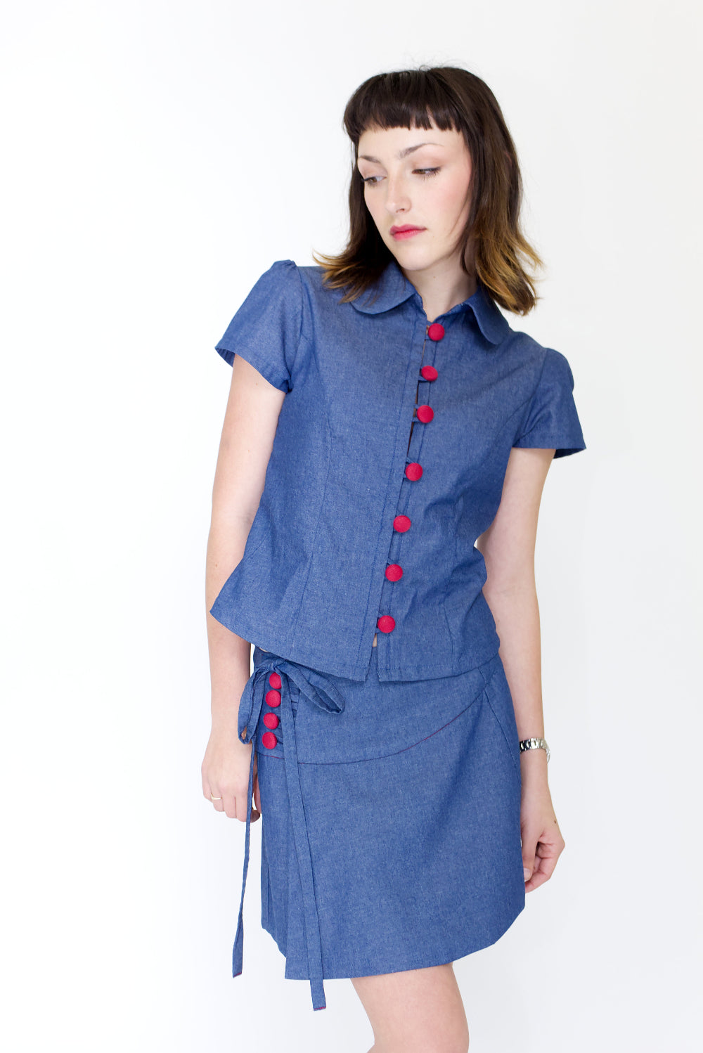 Young woman in a matching blue chambray cap sleeve, peter pan collar, princess line blouse and asymmetrical, low rise mini warp skirt, both with red buttons, in front of a white back ground