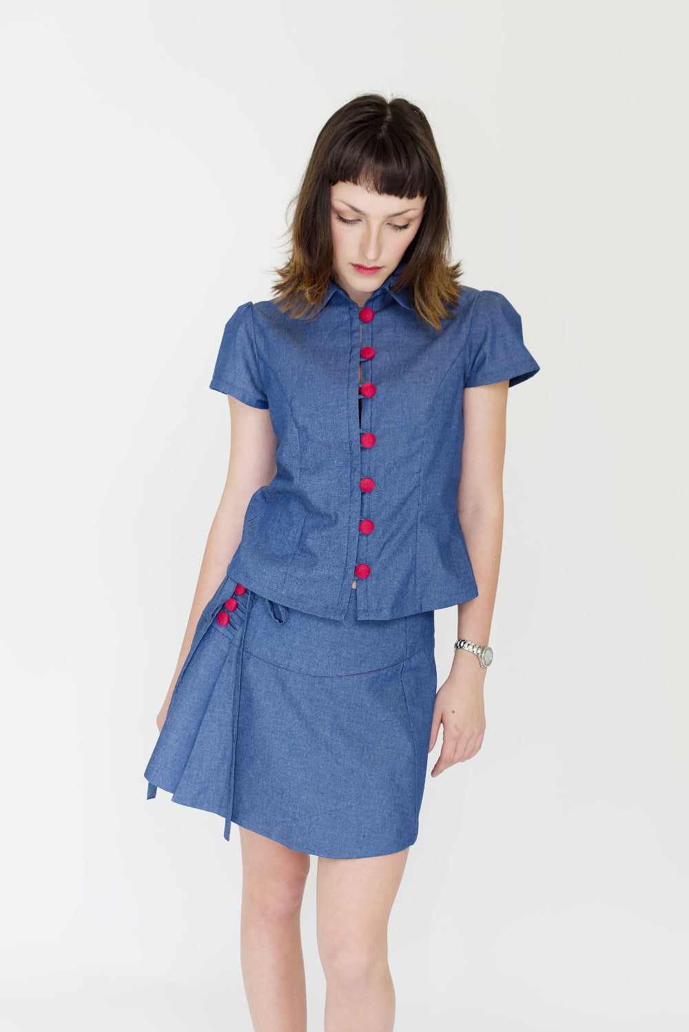 Young woman in a matching blue chambray cap sleeve, peter pan collar, princess line blouse and asymmetrical, low rise mini warp skirt, both with red buttons, in front of a white back ground