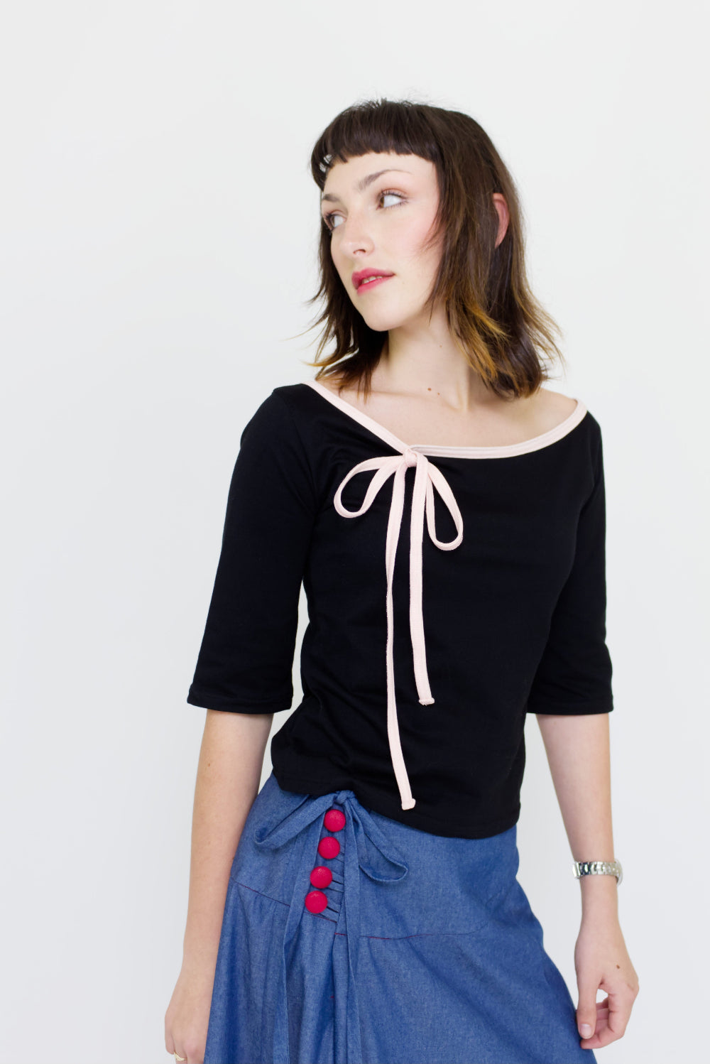 A young woman wearing a black and baby pink boat neck top with a ¾ sleeve and bow, paired with a denim blue chambray maxi skirt, in front of a white back ground